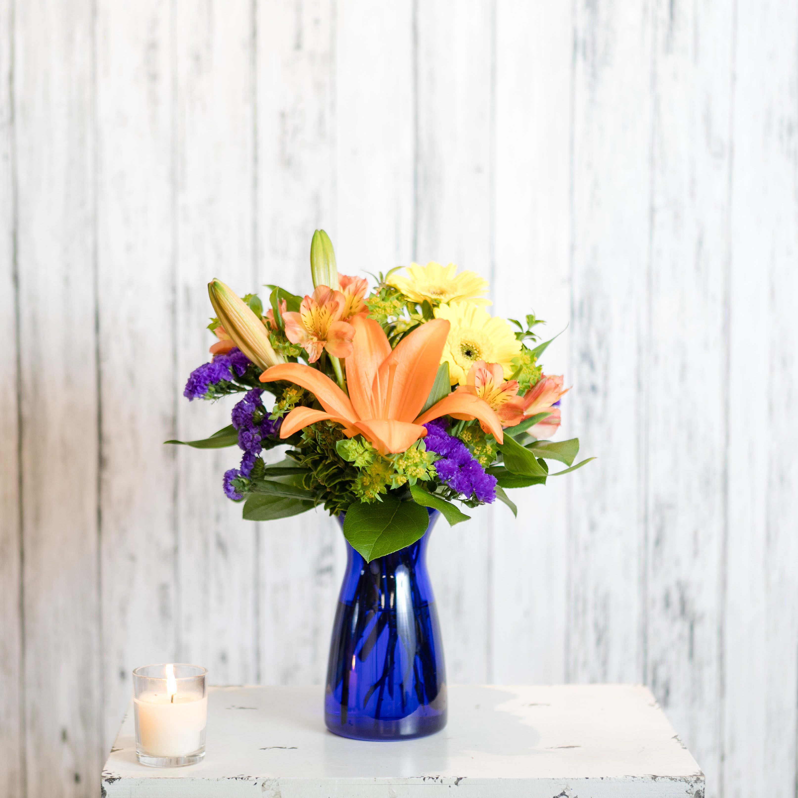Indigo - Orange lilies, alstromeria, yellow gerbera daisies and purple statice combined in a cobalt blue vase will add a bright spot to anyone's day.  Approximate Dimensions: 14&quot; H  x 9&quot; W