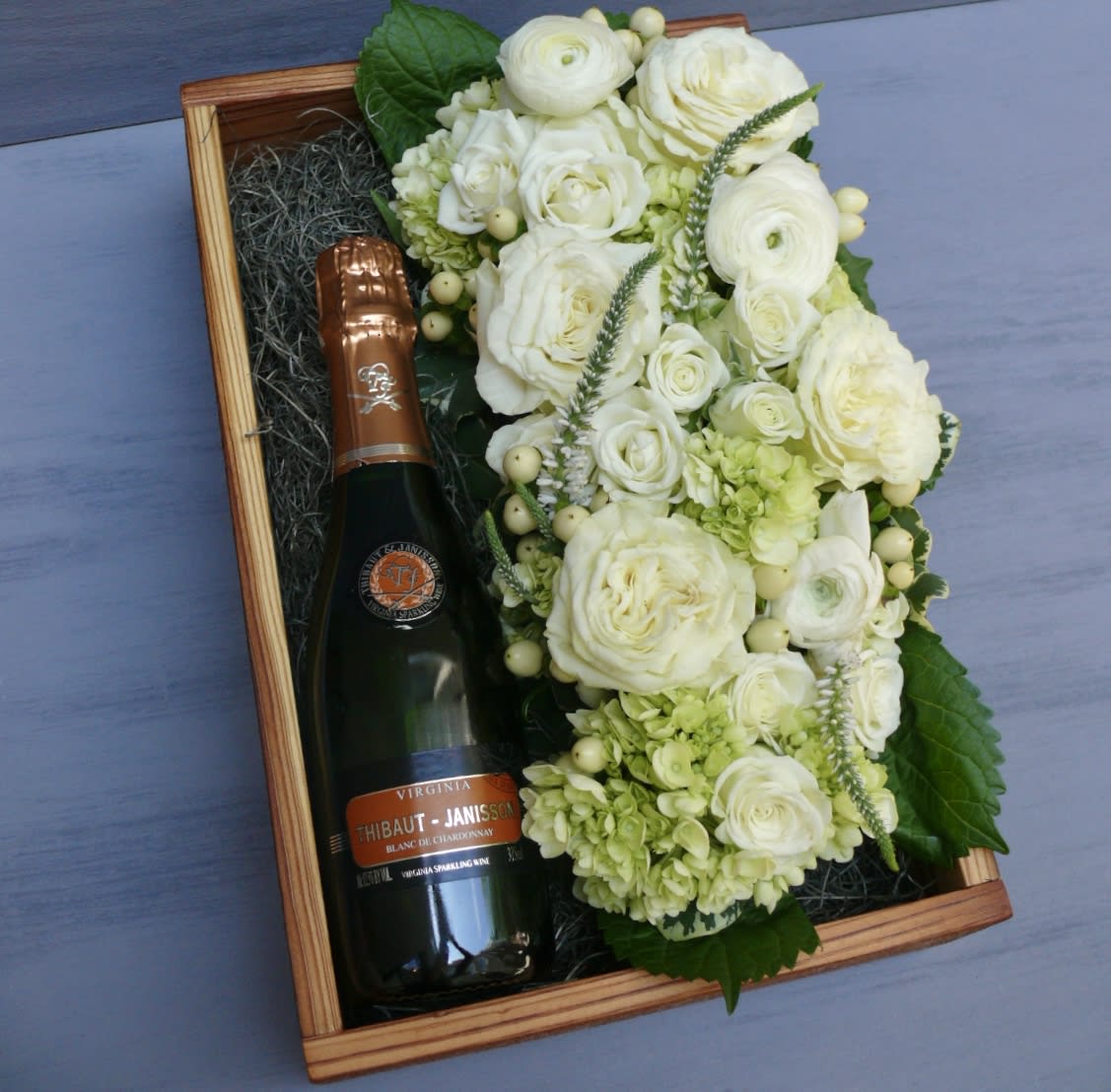 Blanc De Chardonnay Gift Box - A beautiful wooden gift box, locally hand-crafted from reclaimed wood. Thibaut-Janisson's rich yet dry Blanc De Chardonnay rests on a bed of Spanish moss, alongside a beautiful bed of flowers in the pavé style. Your choice of a split 375ml bottle (standard) or a 750ml bottle (deluxe).  You can also specify a color for this arrangement.   Wine Description: This is a truly historic offering, putting Virginia in conversation with the great wines of France’s champagne region. Made from 100% chardonnay grapes, this wine manages to be rich and dry at the same time. This is a bottle made for every kind of celebration. Bubbles go with everything!  Before placing your order, please ensure that the recipient is above 21 years of age and will be home to receive the delivery, as ABC regulation requires that we check photo ID upon delivery.  If no one is present at the delivery site, delivery cannot be completed.  Box measures 8.5x11.5&quot;.