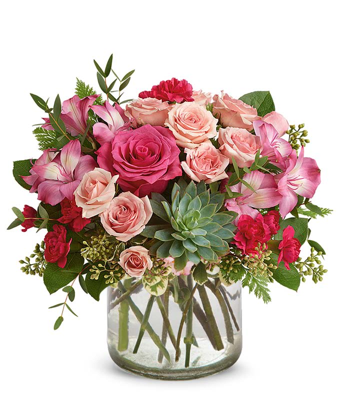 Pink Rose Succulent Garden - Soft, romantic, and effortlessly chic — this floral design brings together classic beauty and modern style. Featuring lush pink and blush roses, delicate alstroemeria, vibrant red mini carnations, and a striking green succulent at the center, this arrangement is a fresh take on timeless elegance. Hints of greenery add texture and balance, all presented in a clear glass cylinder vase for a clean, contemporary finish.  Perfect for birthdays, anniversaries, or simply to brighten someone’s day, this arrangement is a heartfelt gesture wrapped in floral beauty.