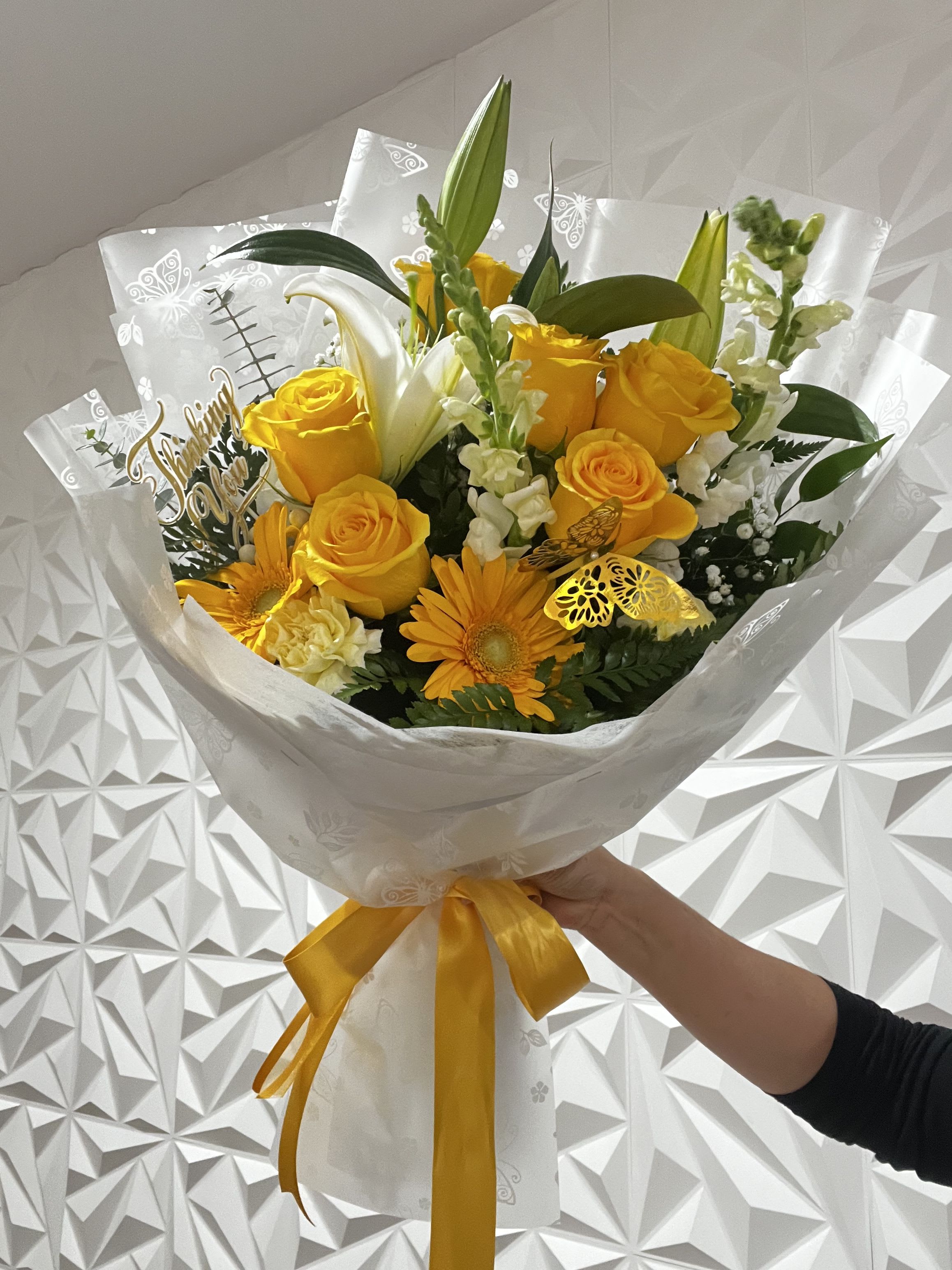 Sunny Elegance - Bright, joyful, and full of meaning, this hand-tied bouquet radiates warmth and positivity. It features 6 vibrant yellow roses, 2 cheerful gerbera daisies, 2 elegant white lilies, 2 graceful snapdragons, and a touch of carnations for added charm and texture. Accented with baby’s breath, baby eucalyptus, and lush greenery, every element is carefully arranged for a perfectly balanced look.  Wrapped in embossed floral paper and finished with a bold yellow satin bow, this bouquet is perfect for birthdays, congratulations, or simply to brighten someone's day with a burst of sunshine.  
