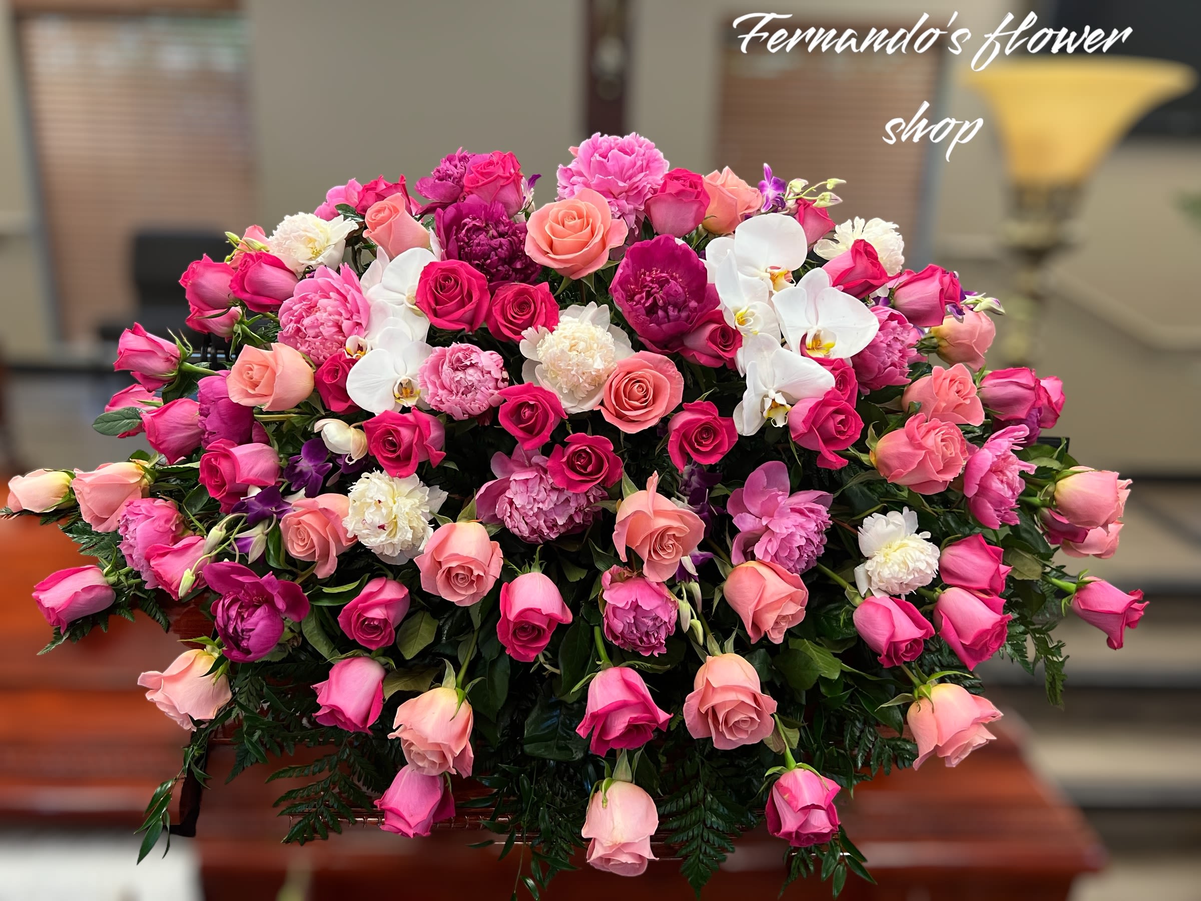 Angelic Light By Fernando’s Flower Shop - The image shows a beautiful floral arrangement atop a polished wooden casket. The arrangement consists of pink roses, peonies, orchids, and other flowers in various shades of pink and white, complemented by lush green foliage. The setting appears to be indoors, likely in a funeral home or memorial service area, with elegant decor in the background, including a brass lamp and wooden blinds.