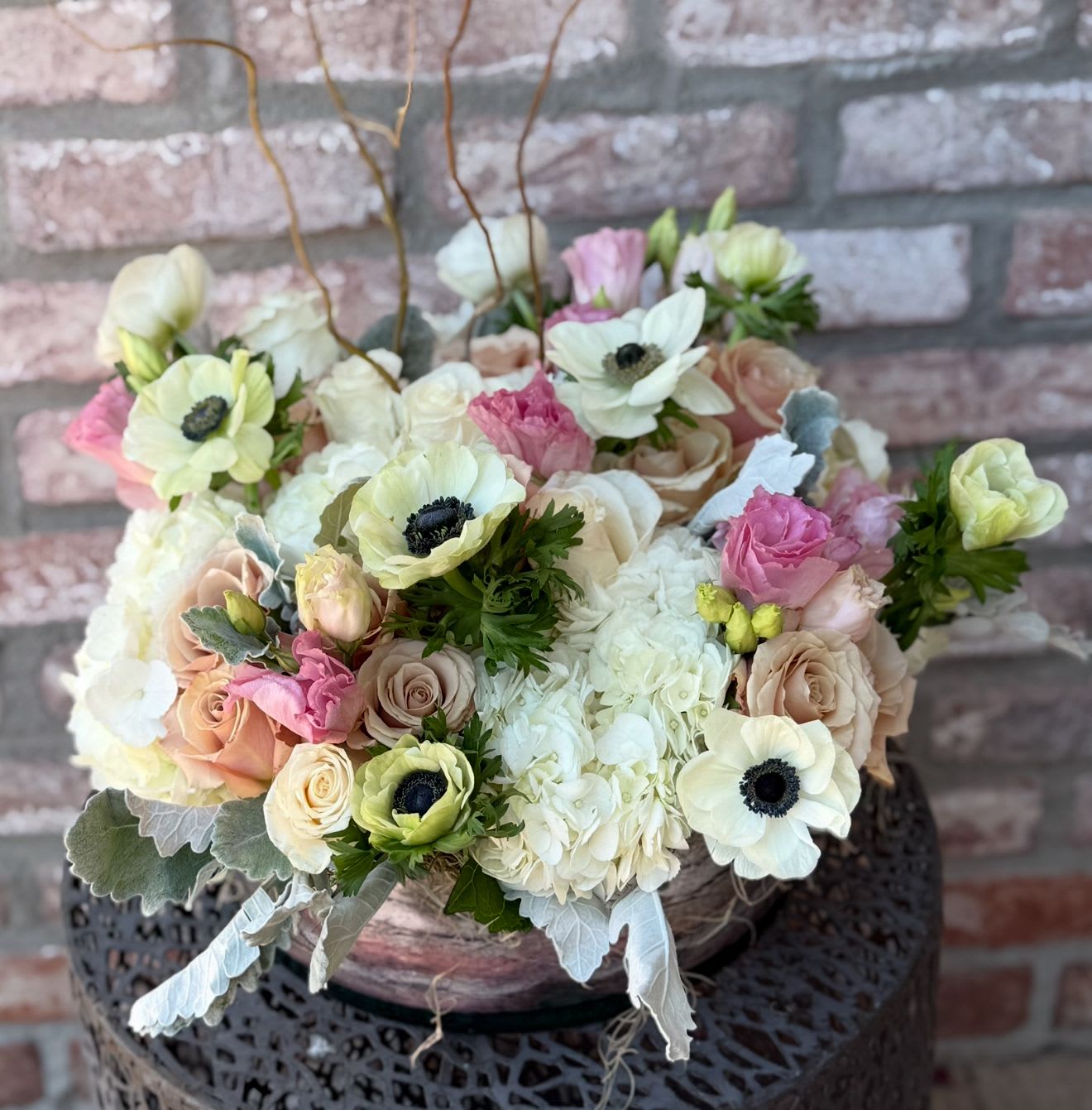 High Society - An arrangement full of sophistication and elegance. Designed in a 12” clear glass dish. Mounded with  fresh lush hydrangea, roses, Veronica, dusty miller and scabiosa pods. Finished off with willow tips. 