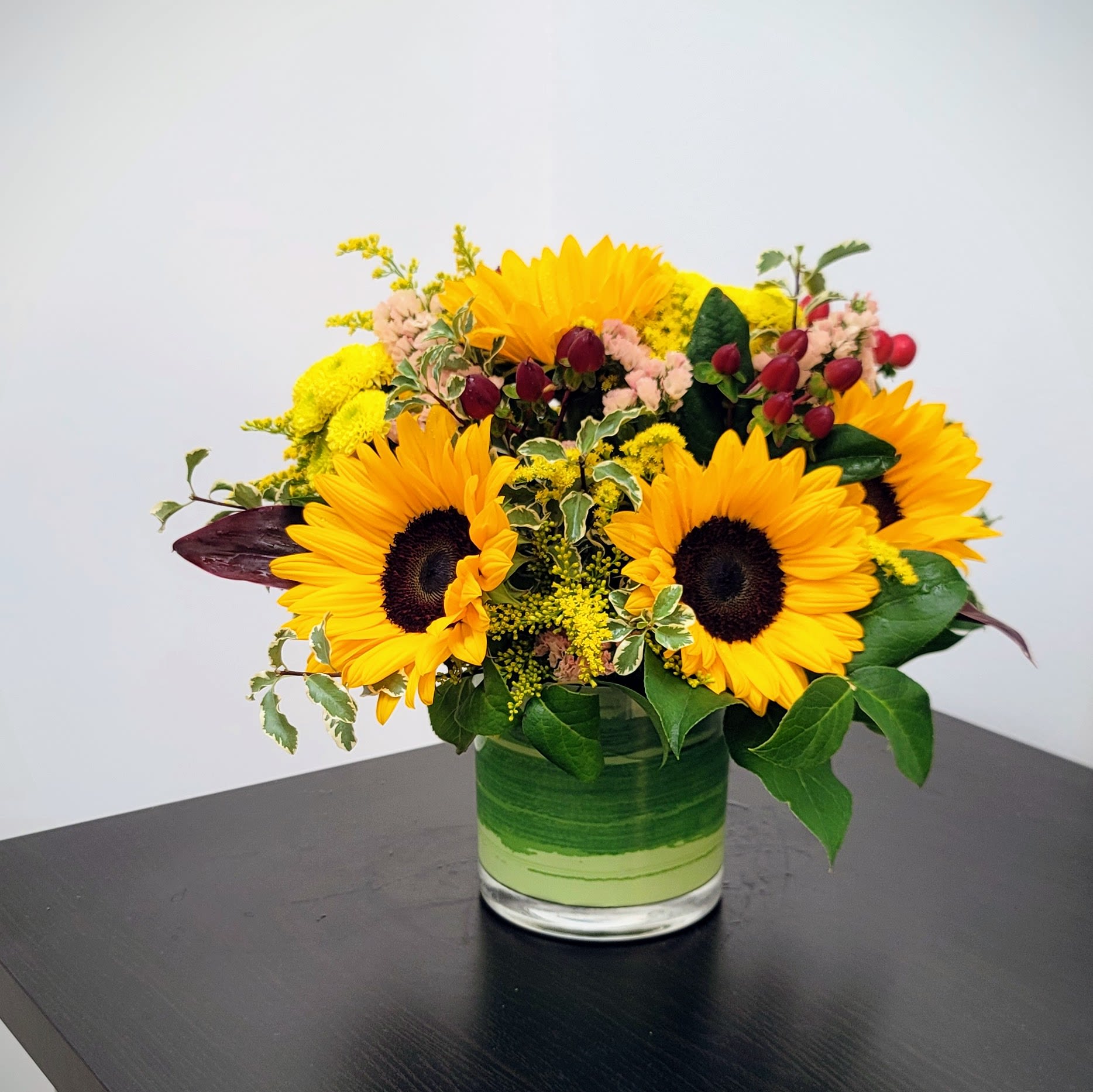 Sunny Sunflowers Bouquet - Bring the warmth of a summer day indoors with our charming &quot;Sunny Sunflowers&quot; arrangement.   This delightful bouquet showcases vibrant sunflowers, beautifully complemented by fresh green foliage and cheerful red berries. Each sunny bloom radiates happiness, making it a perfect pick-me-up for any space.  Approximate Size 12&quot; W x 12.5&quot; H. Brighten their day, order your bouquet today!