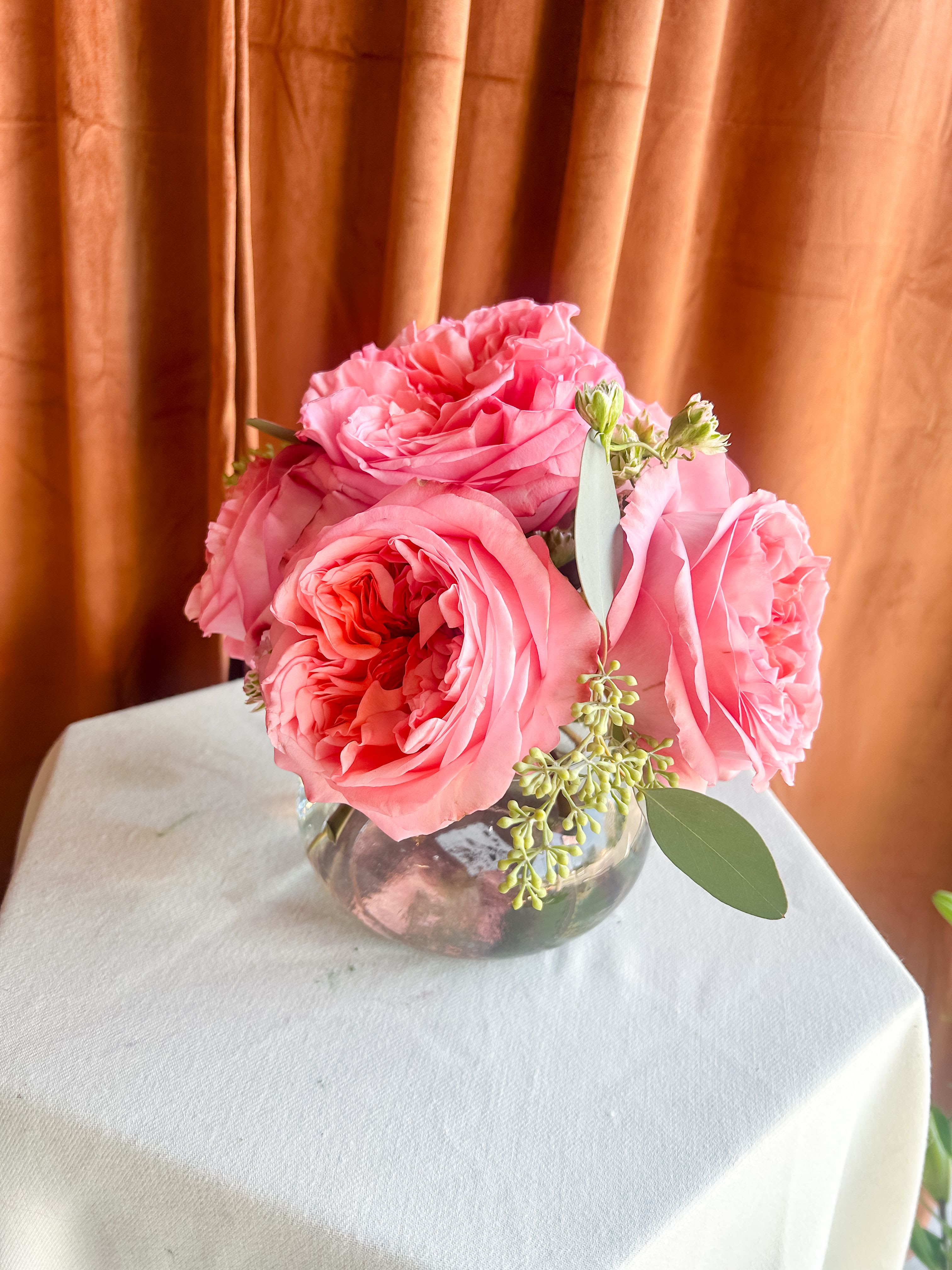 Half Dozen Garden Rose Posey  - A delicate gathering of six lush garden roses arranged in a petite glass vase. Unlike standard roses, garden roses are prized for their captivating fragrance as well as their abundant ruffled petals—making this arrangement as delightful to the senses as it is to the eyes. Accented with soft greenery for a natural touch, this posy is perfect for brightening a desk, side table, or gifting as a simple yet stunning gesture.