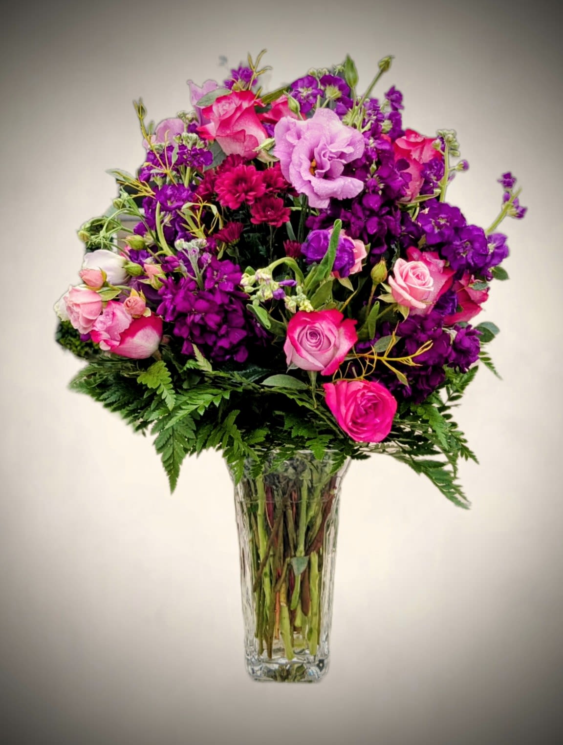 Fuchsia Bliss Fuchsia Bliss - Bold and full of life, our Fuchsia Bliss Vase is a captivating mix of jewel-toned blooms designed to make a statement. Lush hot pink roses, ruffled lavender lisianthus, deep purple stock, and crimson chrysanthemums blend beautifully with soft greenery for a rich, layered look. Arranged in a tall, clear glass vase, this bouquet is the perfect choice for making any occasion unforgettable. Perfect for: Anniversaries, romantic gestures, bold celebrations, just-because surprises Approximate size: 20" H x 14" W