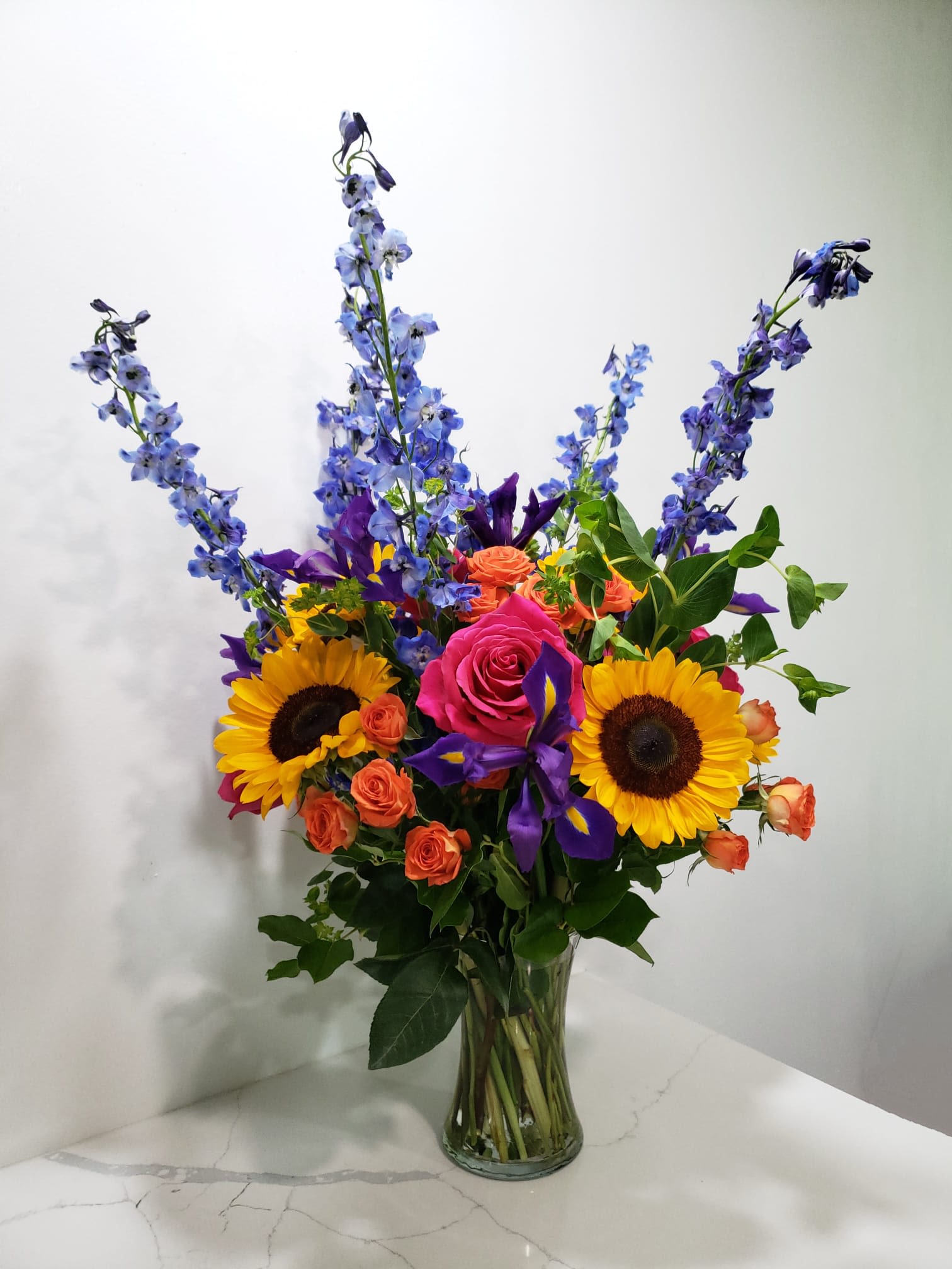 Our Rainbow Our Rainbow - This bright arrangement has blue delphiniums, fuchsia roses, sunflowers, orange spray roses, purple iris and bupleurum in a cylinder glass vase.
