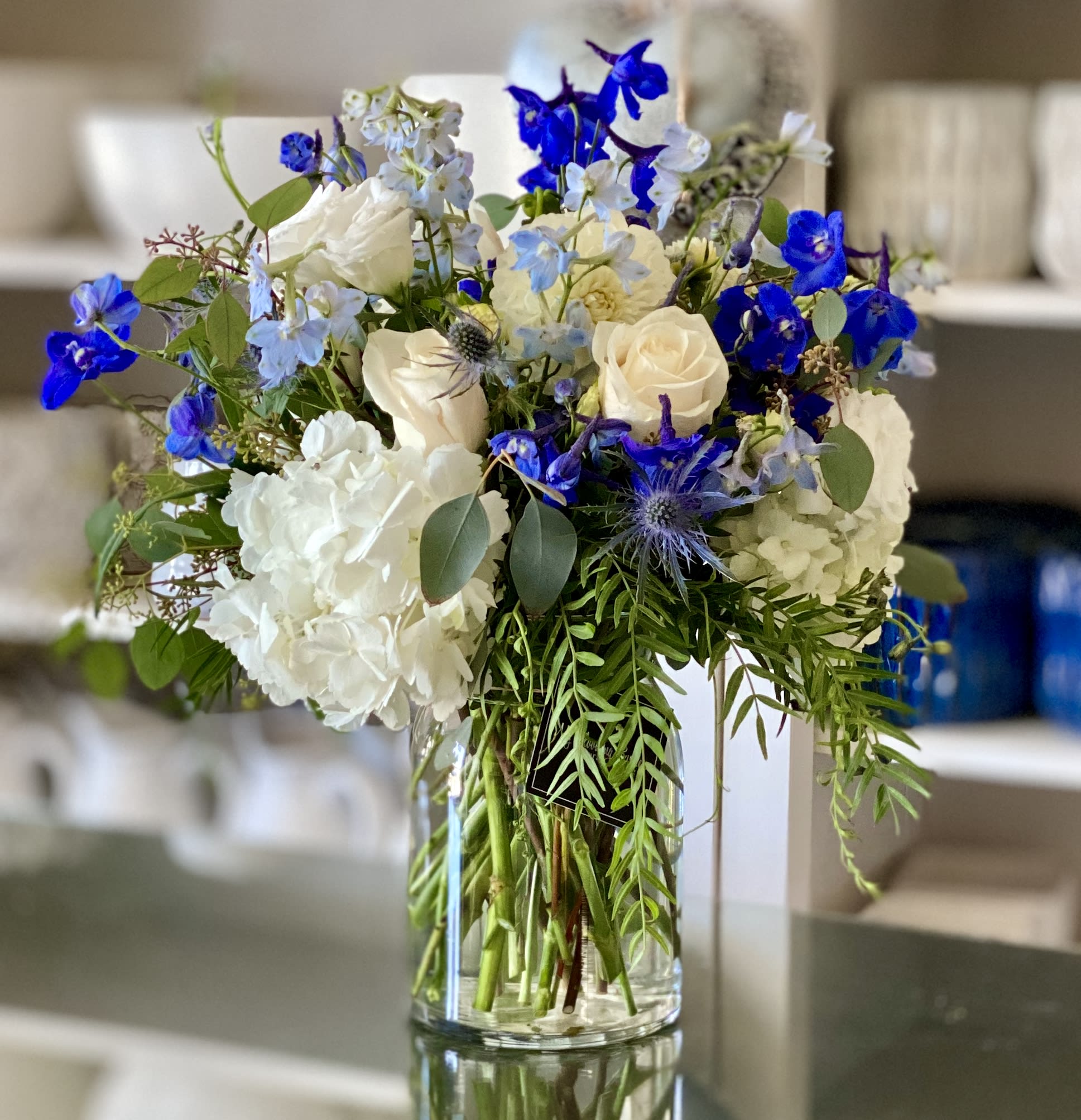 Marina Marina - Are you ready to make a splash at your next occasion? Introducing Marina, our glistening 8' glass vase that's filled with an amazing array of flowers! Enjoy the pure white hydrangeas and roses, expertly complemented by striking dark and light blue delphinium and greenery. Gathering family and freinds around Marina will truly turn heads with its beautiful display! As they say, art is in the eye of the beholder - Marina is an art piece crafted specially for those who want to make a statement on any special night.