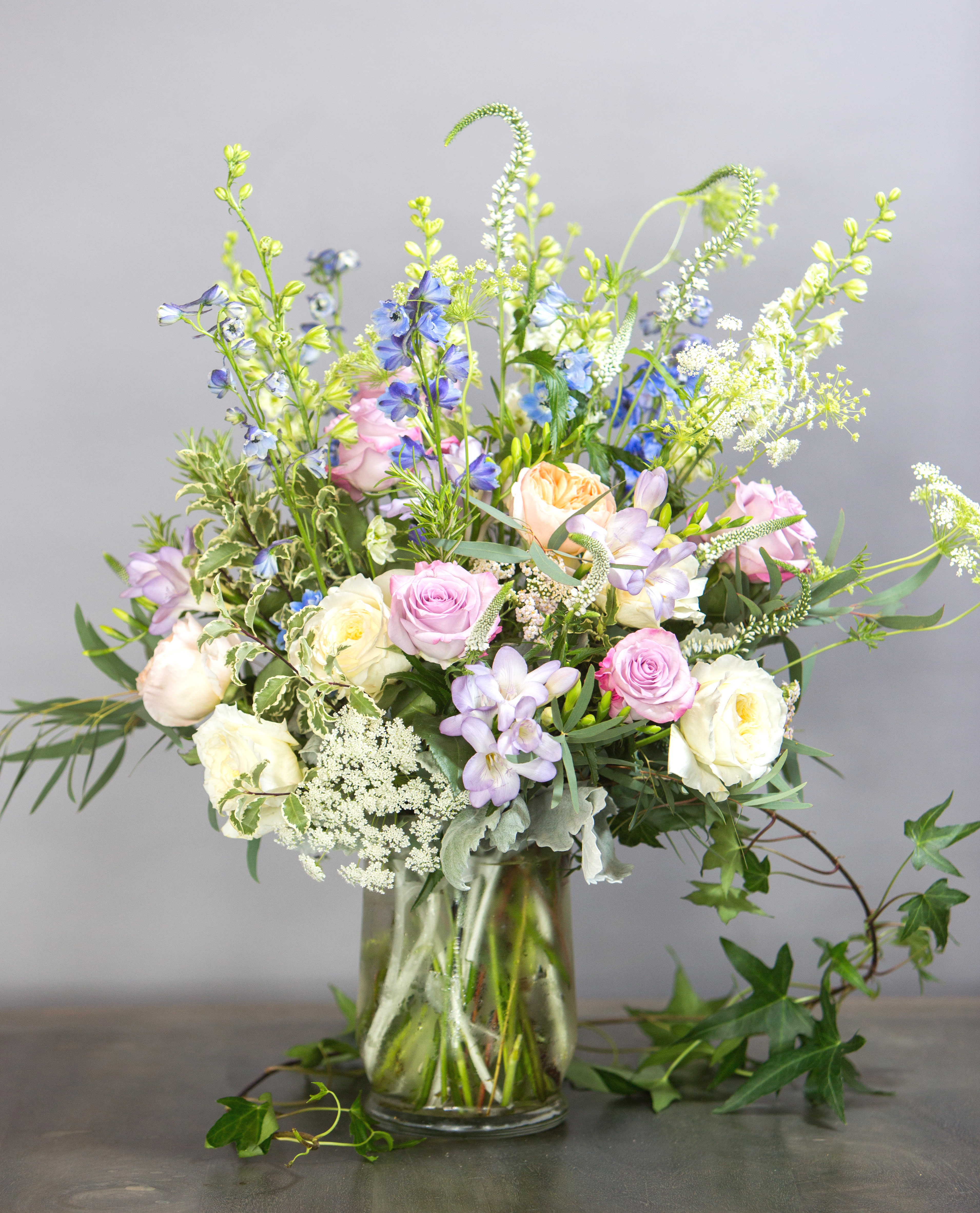 Stone Cottage Collection - Signature Size - A gathering of soft-hued flowers in a grand design inspired by the English countryside. Delphinium, Veronica, Larkspur, Queen Anne's Lace, roses, garden roses and Freesia in shades of lavender, pink, blue and white. Sweet, yet dramatic. Perfect for a birthday, Anniversary or spring occasion.   Arrangement is approximately 16&quot; tall and 10&quot; wide. 
