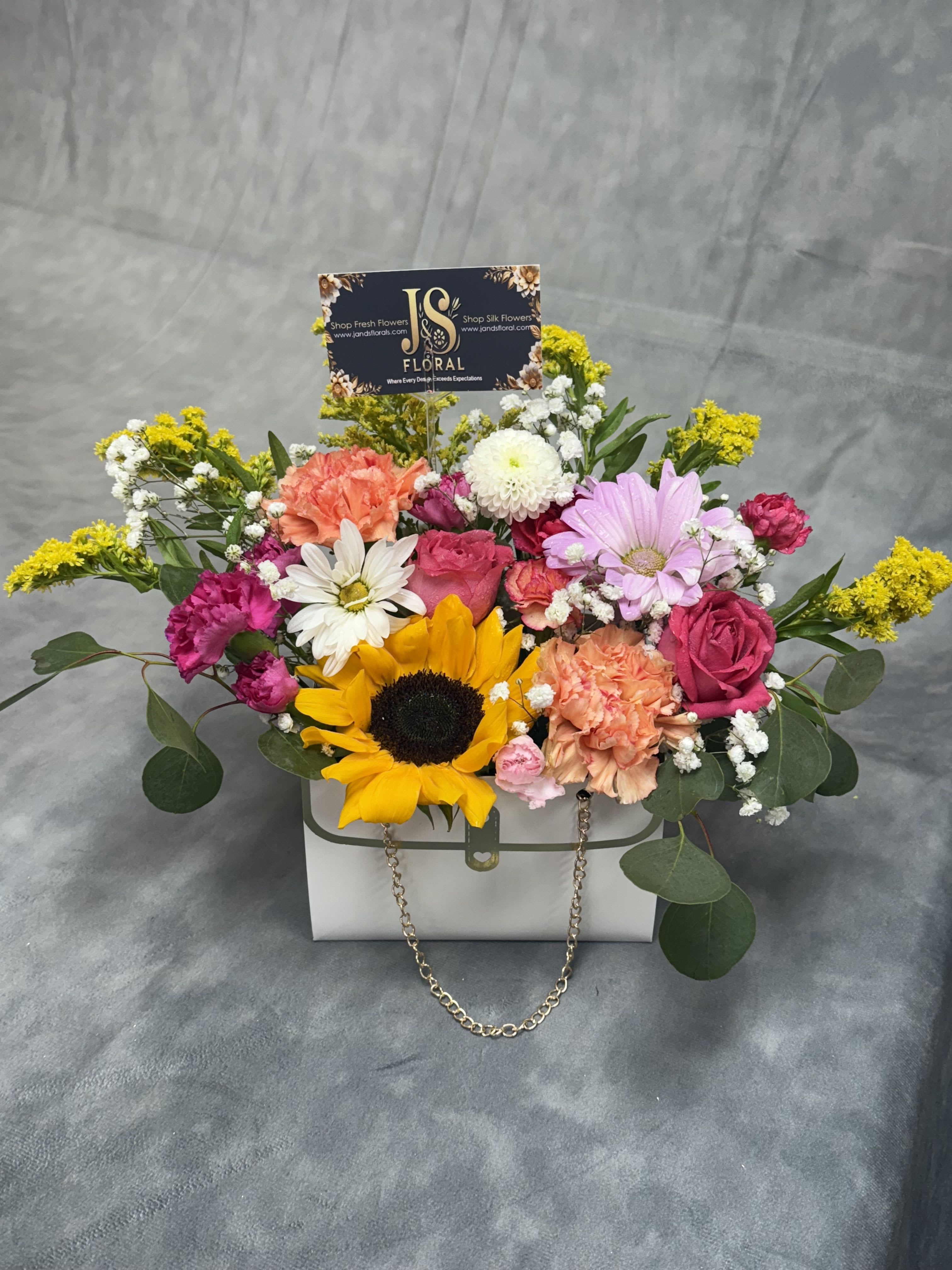 Sunshine Chic Purse  - A charming white purse brimming with a joyful sunflower, vibrant carnations, and playful spray roses in a lively mix of colors — a perfect blend of fun, fashion, and fresh blooms.