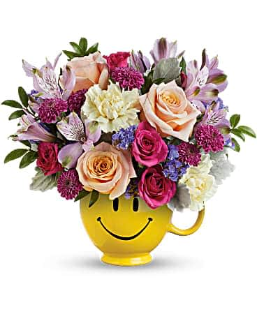 Embrace Happiness Bouquet - Cheer up any occasion with a smile! This fabulously feminine rose bouquet looks extra joyful in a smiling ceramic mug. Peach roses, hot pink spray roses, lavender alstroemeria, light yellow carnations, purple button spray chrysanthemums and lavender sinuata statice are arranged with dusty miller and huckleberry. Delivered in a Be HappyÂ® Mug.