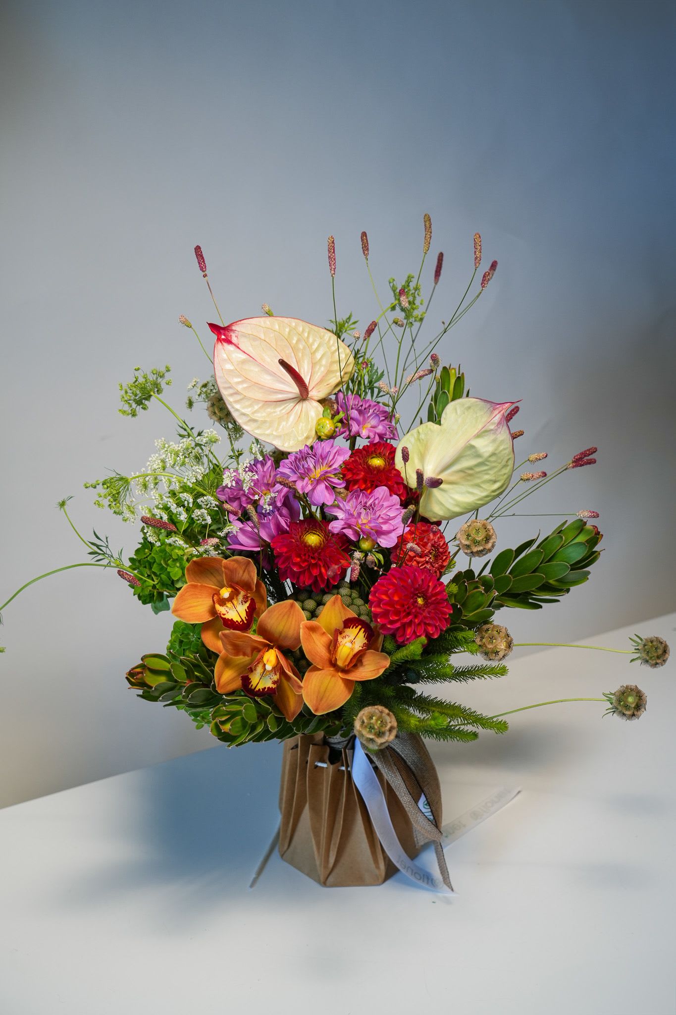 Fire &amp; Ivory Elegance - A bold and sophisticated arrangement blending warm fiery tones with soft ivory accents. Featuring exotic orange cymbidium orchids, creamy anthuriums, rich red dahlias, and vibrant seasonal blooms, complemented by lush greenery and airy wildflower details. Perfect for special occasions or as a striking gift that leaves a lasting impression.