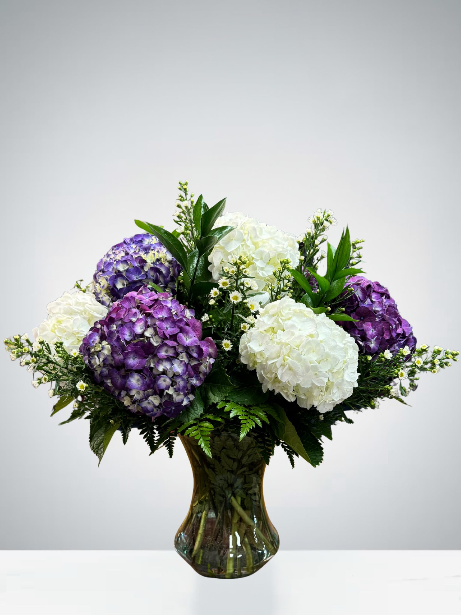 Tranquil Blooms Tranquil Blooms - Graceful and serene, this stunning bouquet features lush purple and white hydrangeas complemented by soft filler florals and rich greenery. Arranged in a classic glass vase, Tranquil Blooms evokes a sense of calm elegance—perfect for expressing sympathy, celebrating a special occasion, or simply brightening someone’s day. The vivid purples add a touch of royalty, while the pristine whites bring purity and peace to any space.