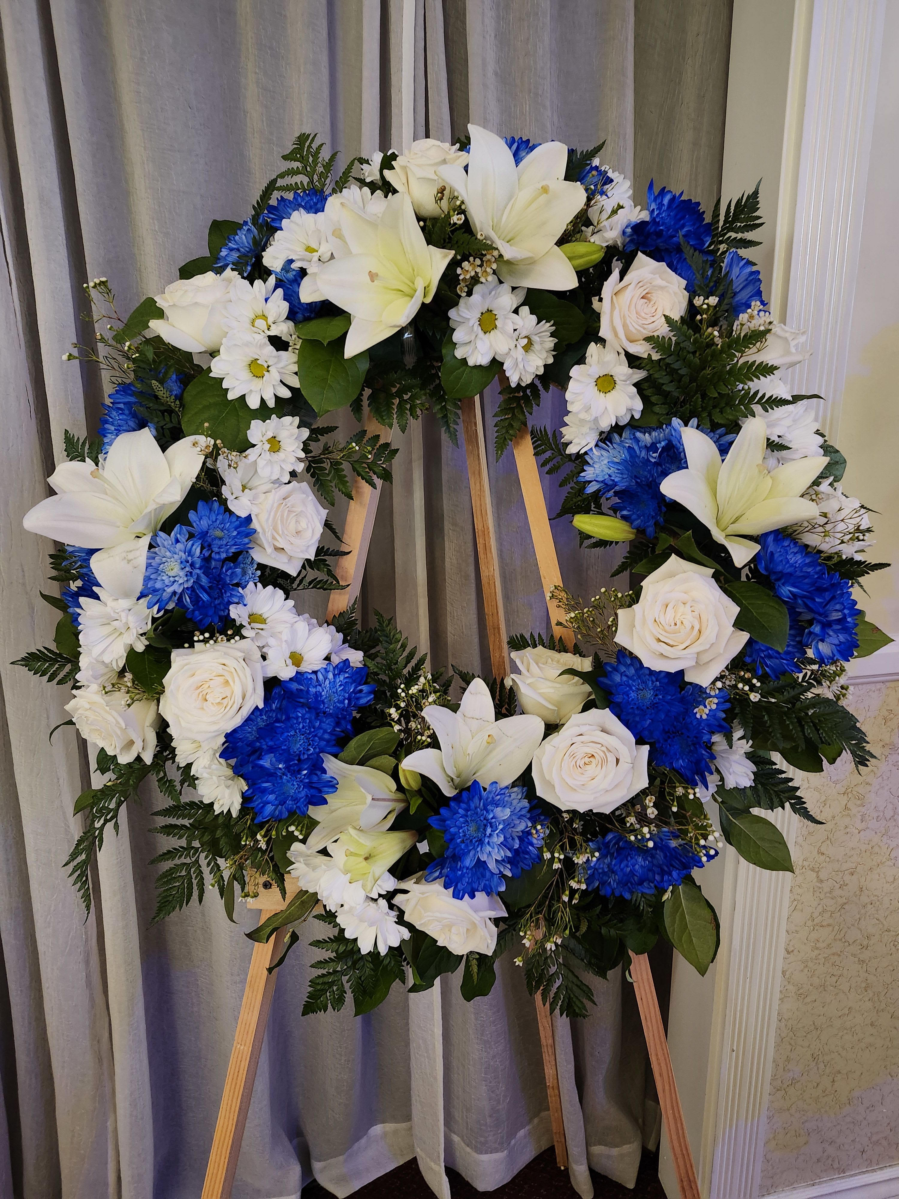 Blue/White Wreath Blue/White Wreath - This wreath is perfect for a male's services. It is designed with roses, lilies, daisies, cushion poms, and wax flowers. Approximate size medium wreath frame of 18-20 inches wide, with floral additional 3-4 inches more. The banner can be purchased by searching "Sympathy Banner"