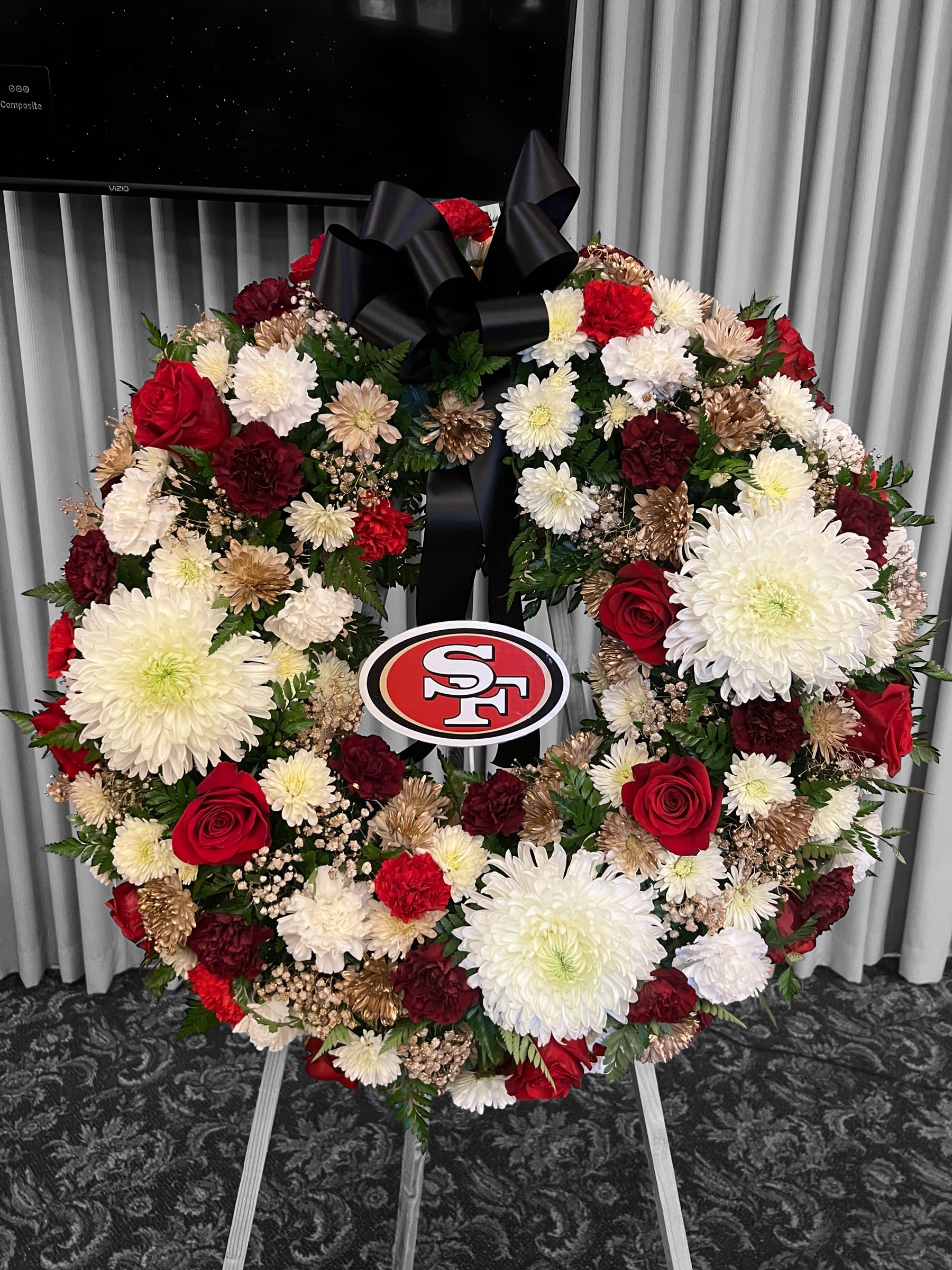 49 Wreath By Fernando's Flower Shop - “A heartfelt and personalized funeral wreath designed to honor a cherished life. This circular arrangement features an elegant mix of white and red flowers, including large white chrysanthemums symbolizing peace and love, alongside rich red roses and carnations for respect and remembrance. Golden floral accents add a touch of warmth and grace, while a prominent black ribbon conveys solemnity and reverence. At the center, the San Francisco 49ers logo pays tribute to a devoted fan, making this wreath a truly meaningful and unique expression of love and loss. ️” STANDARD SIZE 31 INCHES
