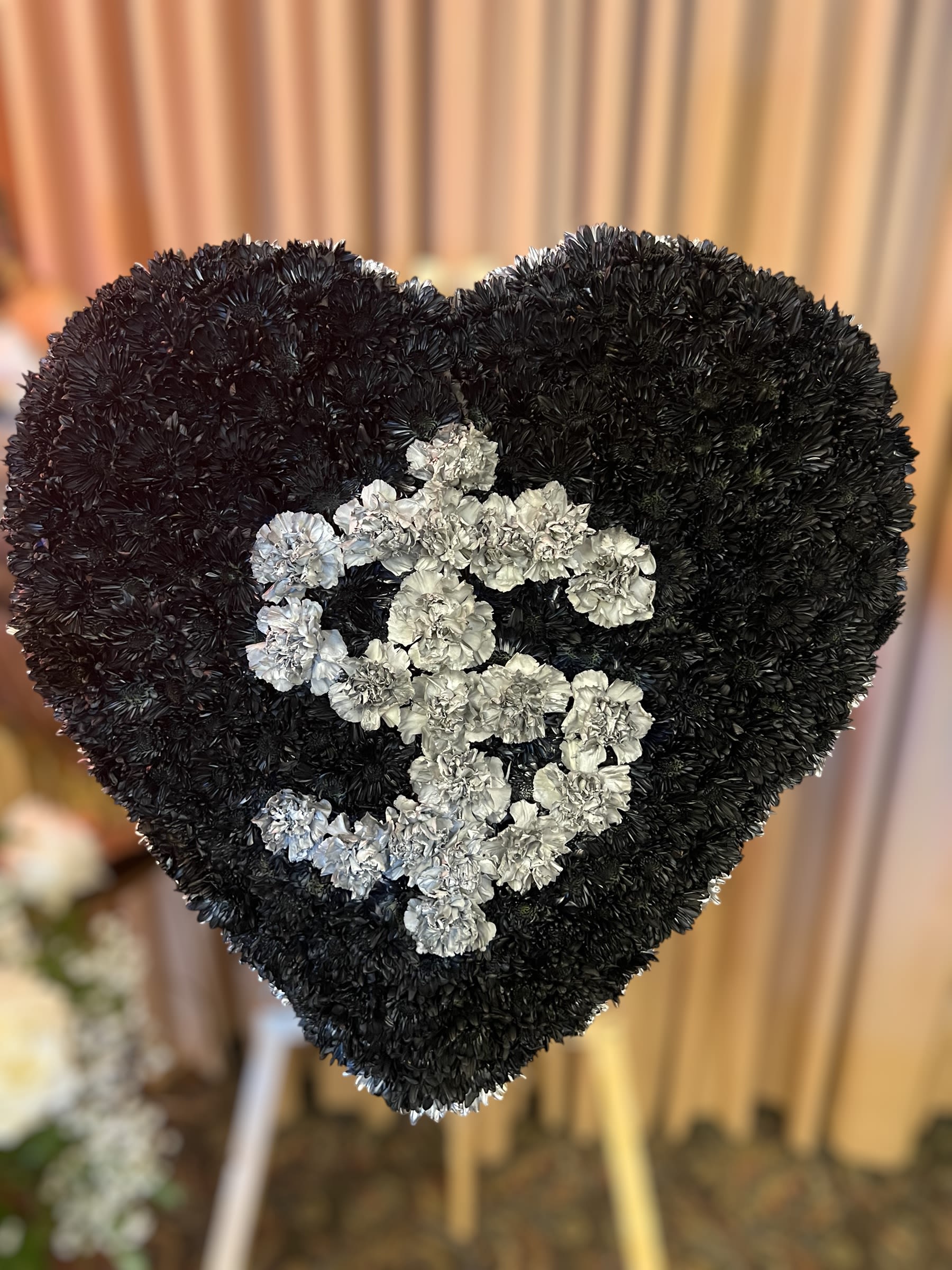 Black Heart - This unique funeral arrangement features a heart-shaped design crafted from black carnations, symbolizing deep sorrow and remembrance. At the center of the heart, a striking silver dollar sign made from white carnations stands out, representing a personal or professional connection to finance or wealth. This custom display is a meaningful tribute to the individuality and legacy of the departed, offering a bold and heartfelt statement. STANDARD SIZE 30 INCHES