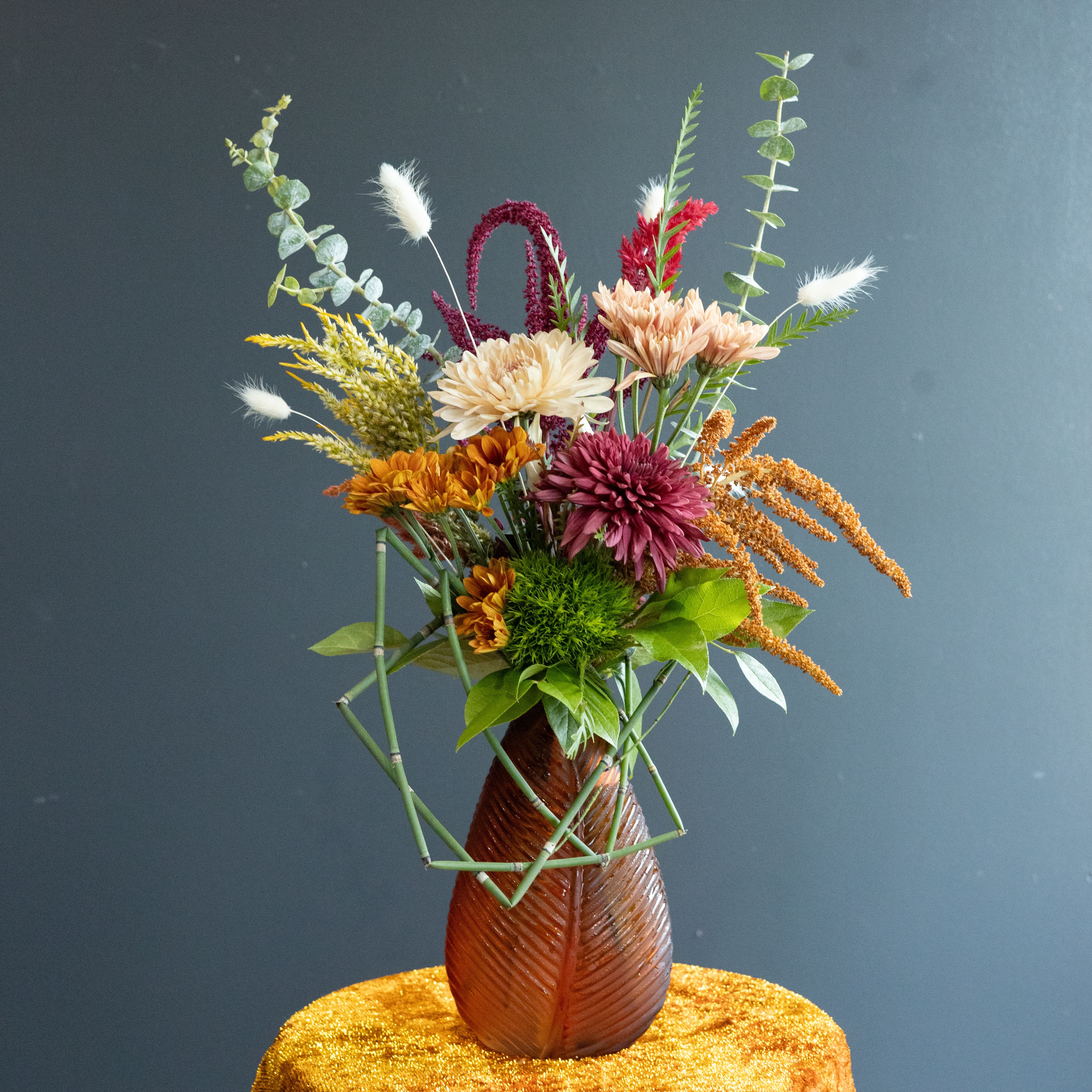 Crimson Bonfire - Gorgeous splashes of crimson colored florals in a fiery hot floral design with artistic bamboo touches in a autumn colored glass vase.
