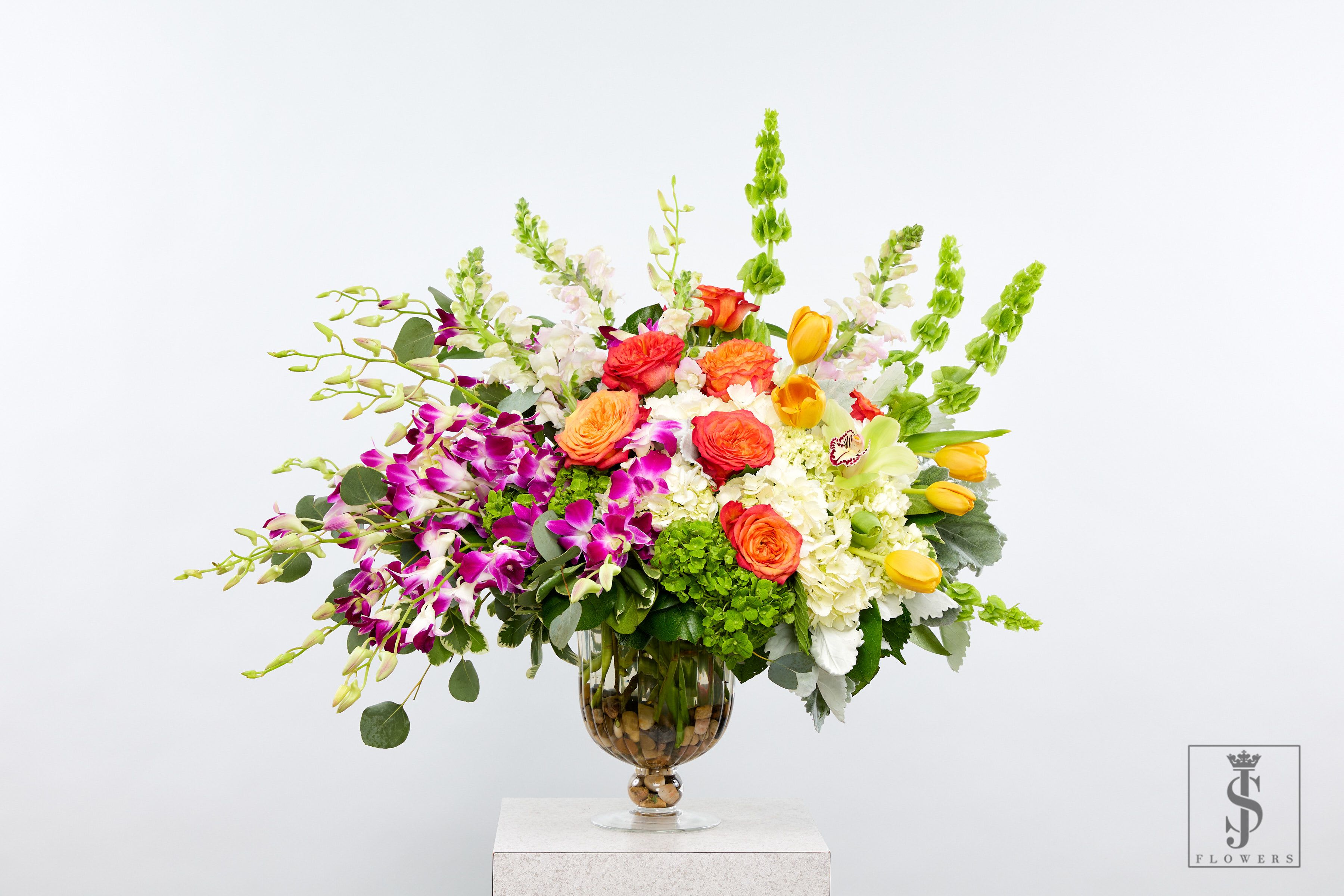 Royal Radiance - Make a show-stopping impression with our luxury oversized floral arrangement, bursting with vibrant tropical blooms, rich textures, and elegant layers. Designed with orange roses, yellow tulips, purple orchids, cymbidium orchids, and more, this lush piece is a floral celebration in every sense.  Arranged in a tall clear urn vase with decorative stones, this premium bouquet is perfect for weddings, large events, hotel lobbies, luxury home staging, and milestone celebrations. A true statement piece available for premium delivery across the entire DFW area — impress with color, scale, and class.