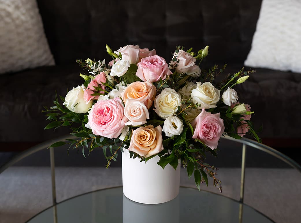 Blush Collection - Signature Size  - Shades of pink, soft peach and ivory reminiscent of a vintage English garden come together to make our Blush Collection. Flowers include garden roses, roses, Lisianthius and white hydrangea with accents of Italian Ruscus and Eucalyptus.   Our J. Morris Signature designs are perfect for an anniversary, birthday, as a thank you or as a larger centerpiece for a kitchen island or table. They are low enough to see over but are still full and grand. Featured in a white 6” ceramic vase this arrangement is 15” high and 12 inches wide.  
