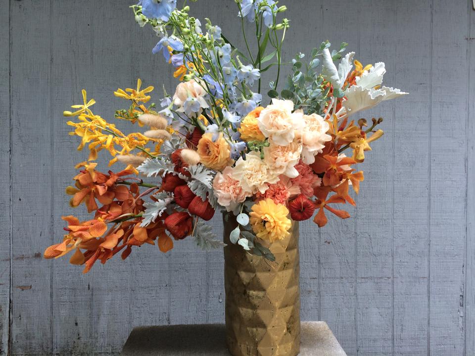 Shades of Fall Shades of Fall - Fun, cheerful, and perfect for the end of summer, this medium-tall arrangement in stunning dusty sky blues, oranges, and yellows is sure to make someone happy. Light blue delphinium is sprinkled with the gray of dusty miller and hints of dried grasses, along with jack o'lantern pods, blush French carnations, and mokara orchids, all arranged in a modern, geometric gold ceramic vase.