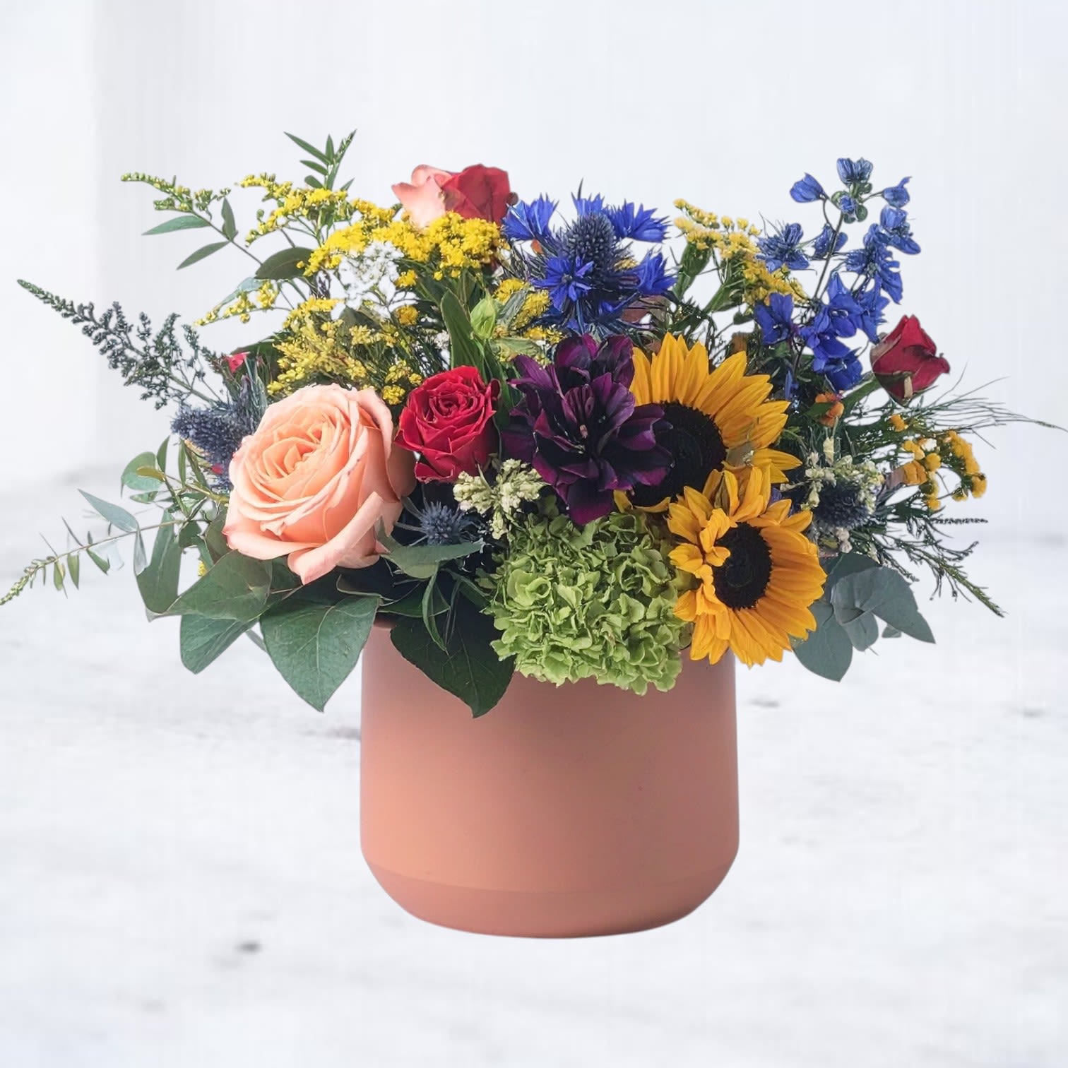 Cheerful Garden Drop Cheerful Garden Drop - Capture the essence of a sunlit garden with Cheerful Garden Drop, a vibrant floral arrangement that radiates warmth and joy. Designed in a chic matte terracotta vase, this breathtaking bouquet features golden sunflowers, peach garden roses, deep blue delphinium, and delicate yellow alstroemeria, accented with lush greenery and textural elements for a naturally elegant touch. Perfect for birthdays, celebrations, or simply brightening someone's day, Cheerful Garden Drop brings a lively and organic beauty to any space. Available for same-day delivery in Houston and surrounding areas, this arrangement is a stunning way to share happiness and love.