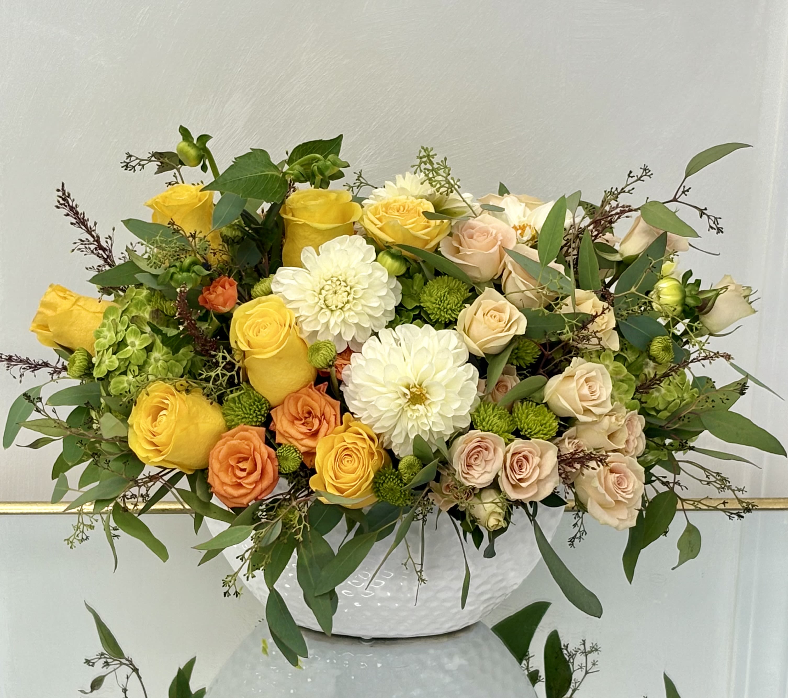 Golden Harvest By VLM Flowers - A radiant medley of seasonal beauty, Golden Harvest combines sunny yellow and soft peach roses, white dahlias, green hydrangea, and playful pops of orange. Set in an elegant textured white ceramic bowl and framed by lush greenery, this arrangement evokes a feeling of warmth, gratitude, and natural charm—ideal for home décor, thank-you gifts, or autumn celebrations.