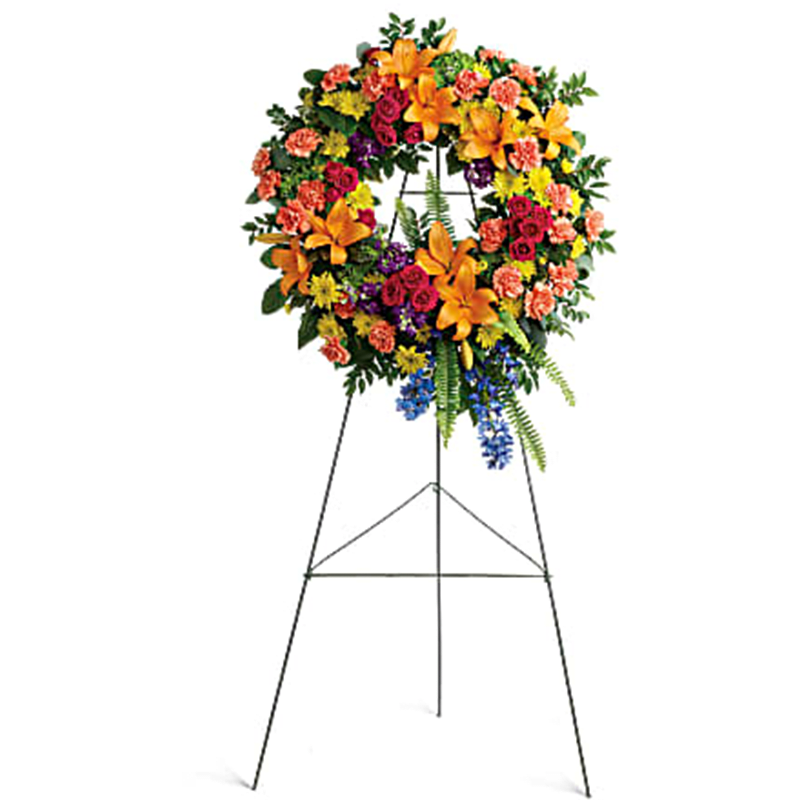 The Colorful Serenity Wreath Standing Spray - Like a beacon of love and hope, this glorious wreath of hydrangea, roses and lilies echoes the joy of your precious memories. This wreath features green hydrangea, hot pink spray roses, orange asiatic lilies, orange carnations, blue delphinium, purple stock, yellow cushion spray chrysanthemums, huckleberry, variegated pittosporum, sword fern, silver dollar eucalyptus, and lemon leaf. Delivered on a wire easel.