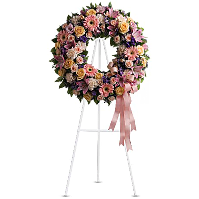 The Graceful Wreath Standing Spray - Family and friends will recollect how special their loved one was with this gentle ring of pastel blooms. Highlighted with a pink satin ribbon, it is a lovely reminder of sweet memories and beautiful times shared. Peach roses, gerberas and carnations, pink asiatic lilies and lisianthus, along with purple limonium and lavender larkspur - accented by greens woven throughout - are presented on an easel-mounted wreath, ideal for a memorial occasion. Orientation: One-Sided