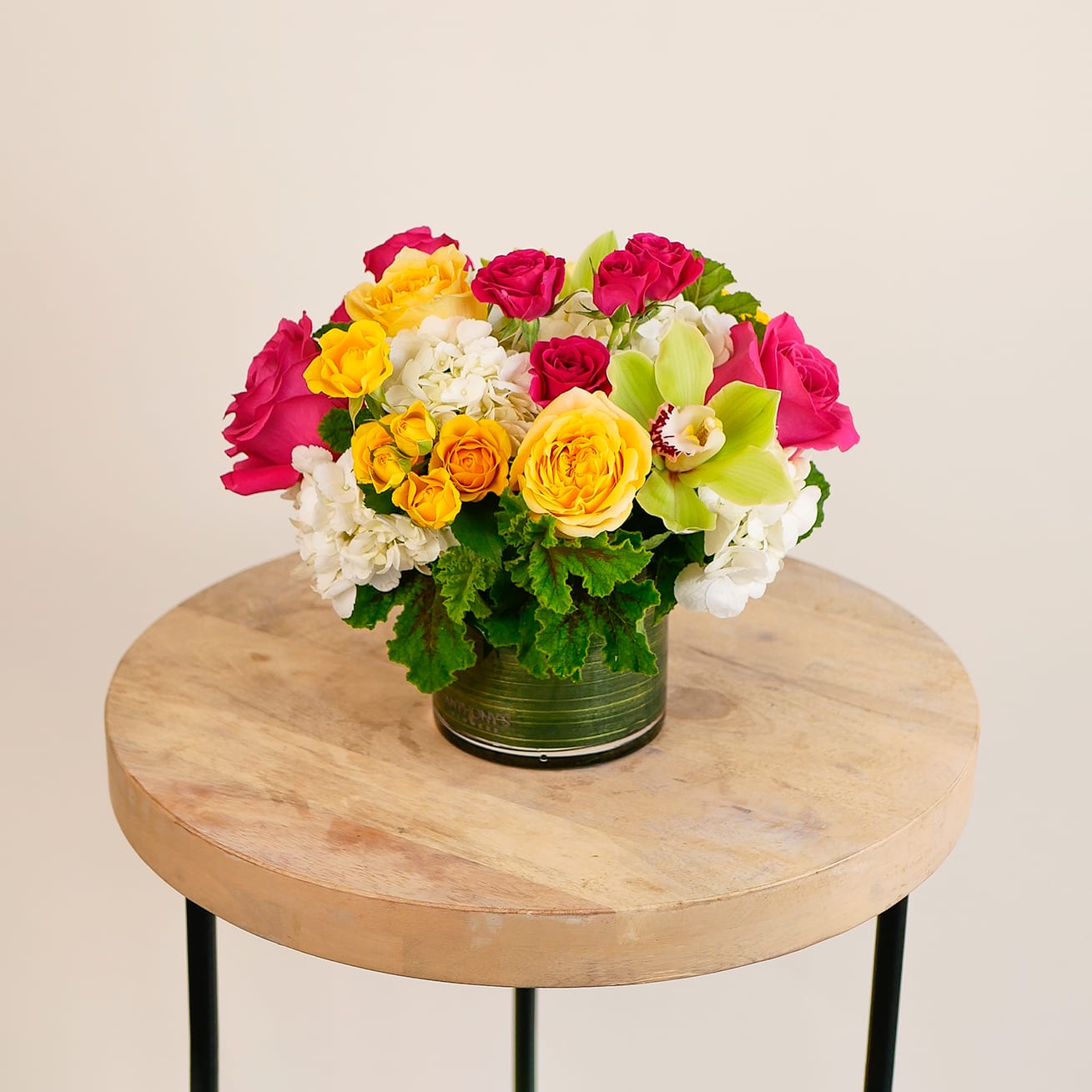 Uptown Girl - This colorful arrangement features a fun collection of white, yellow and hot pink blooms, such as roses, spray roses, hydrangeas and cymbidium orchids in a clean leaf-lined glass cylinder. Upgrade to the Deluxe to add premium pink double tulips and seasonal ranunculus in a larger 6x6&quot; vase.
