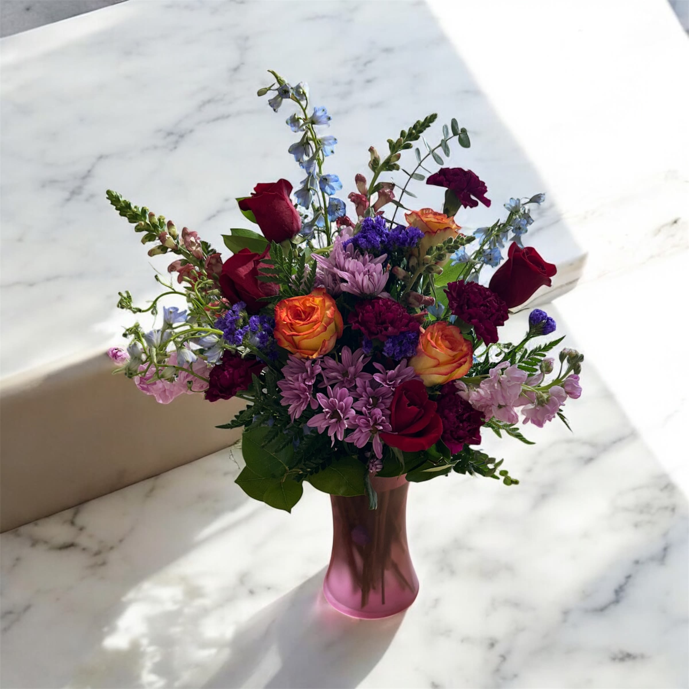 Rainbow Romance Bouquet - A stunning mix of blooms in bold and soft hues, beautifully arranged in a charming pink vase. Towering blue delphiniums pair with passionate red and bright orange roses, while soft pink stock and snapdragons add texture and charm. Lavender daisies and rich purple carnations complete the arrangement, creating a vibrant, elegant display perfect for celebrating love, joy, and life’s special moments