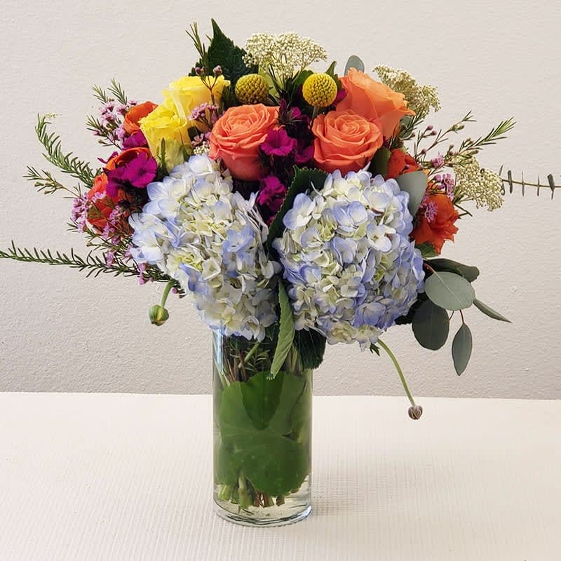 Amaze Bouquet  - A vibrant mix of yellow and orange roses beautifully contrasted with blue hydrangeas and ranunculus, accented by purple mini carnations.   Arranged in a sleek cylindrical glass vase, this stunning bouquet is perfect for Mother's Day, birthdays, anniversaries, or any special occasion.  Approximate Size: 17&quot; H x 15&quot; W. Order now for fresh flower delivery!