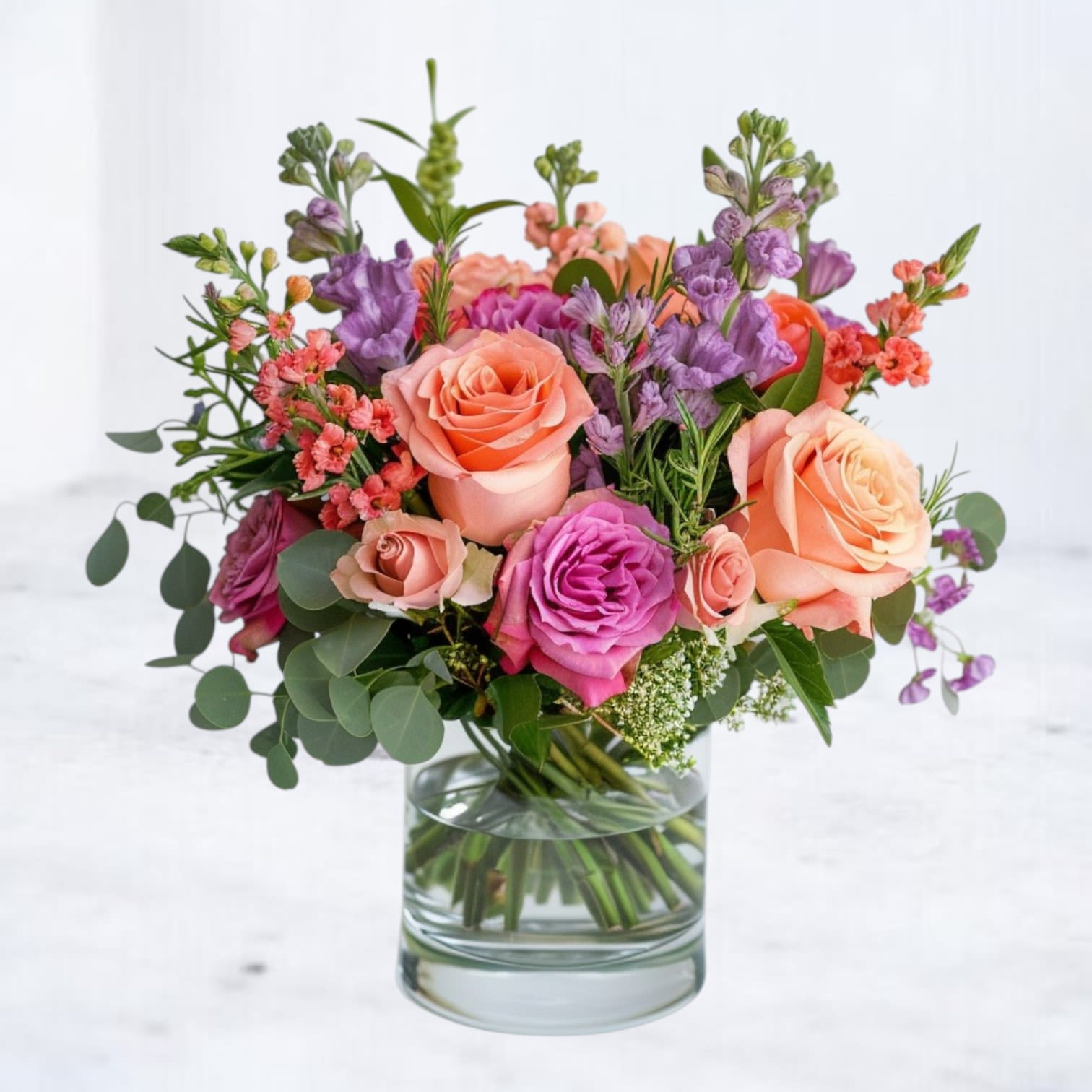 Meadow Charm - Bright, cheerful, and artfully arranged, the Meadow Charm bouquet brings a burst of modern garden beauty to any space. Featuring a harmonious blend of vibrant coral roses, soft peach blooms, vivid fuchsia spray roses, delicate purple stock, and airy accents of seeded eucalyptus, this lush arrangement feels freshly gathered from a blooming meadow. Its loose, airy design and layered textures make it the perfect gift for birthdays, anniversaries, thank-you gestures, or simply brightening someone’s day. Designed with a modern twist on classic floral beauty, the Sunset Meadow Charm is hand-delivered in a stylish 4x4 glass vase for a polished, ready-to-enjoy display.    modern garden flower arrangement coral and purple roses bouquet Magnolia TX flower delivery
