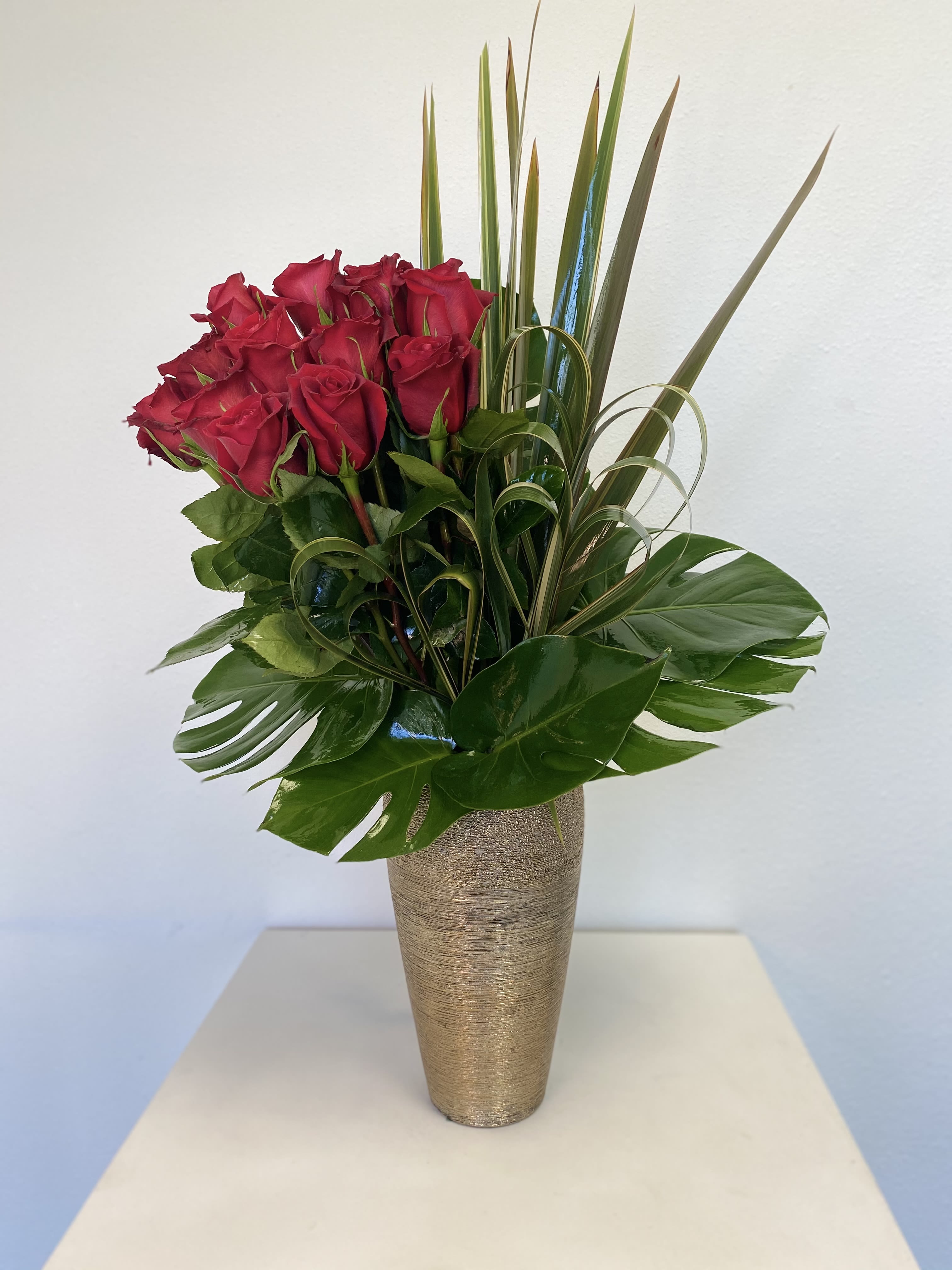 Valentina - Make a bold statement with this striking contemporary arrangement that combines passionate romance with exotic sophistication. This stunning design features a dozen premium red roses arranged in a tight, luxurious cluster, symbolizing timeless love and devotion.  The dramatic floral display is elevated by an artful selection of tropical foliage including broad monstera leaves with their signature splits, and spiky flax leaves that creates striking vertical lines. Adding a romantic touch, select tropical leaves have been carefully shaped into heart forms, creating a playful yet sophisticated detail that enhances the love theme. This exotic greenery transforms a classic rose arrangement into a modern masterpiece with architectural appeal and romantic symbolism.  Presented in a sophisticated textured gold vessel with contemporary metallic finish, this arrangement brings glamour and drama to any setting. The warm golden tones beautifully complement the rich red roses while the sleek tapered silhouette adds modern elegance.  Perfect for: - Romantic anniversaries and Valentine's Day - Modern home and office décor - Contemporary celebrations and milestone events - Corporate gifting with sophisticated appeal - Date nights and intimate dinners - Anyone who appreciates bold, dramatic design with romantic details  Approximate dimensions: 14&quot; wide x 28&quot; tall