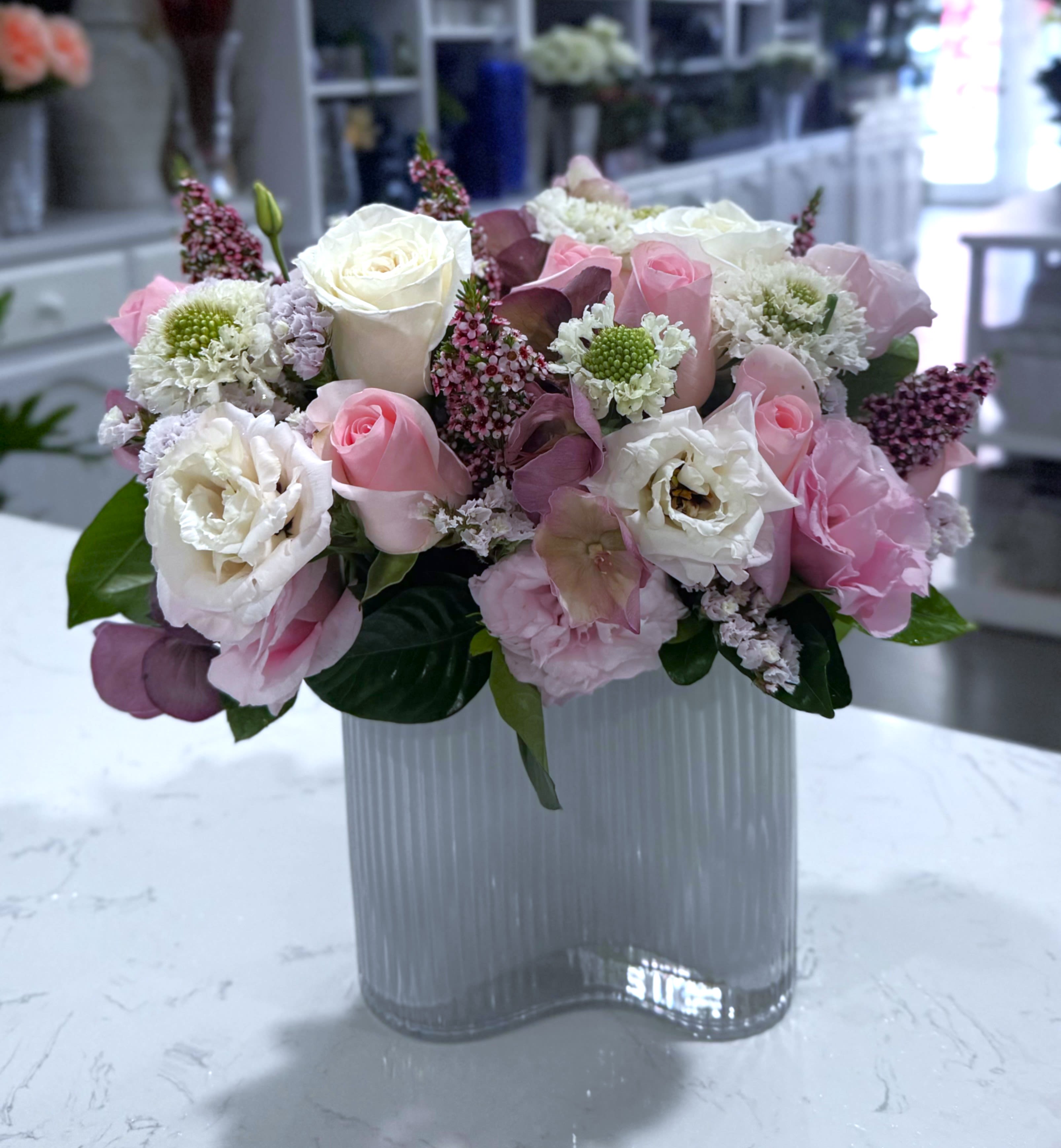 Velvet Blooms - A lush medley of blush roses, lisianthus, scabiosa, and soft textural accents in rich mauves and creamy whites. Designed in a sculptural wave vase, Velvet Bloom is effortlessly modern with a romantic twist—ideal for celebrating love, beauty, and everything in between.