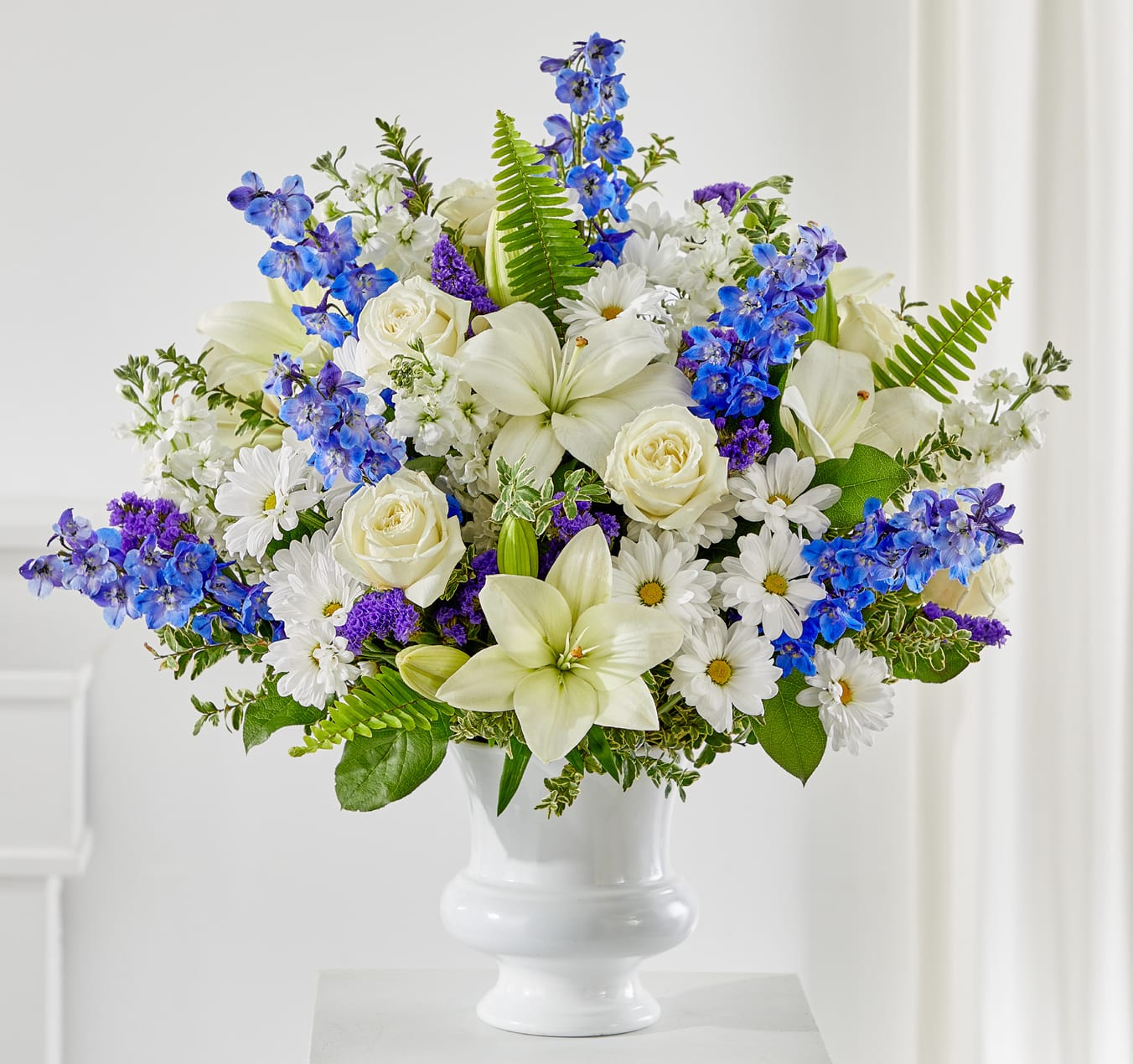Expressions of Hope Arrangement - The Expressions of Hope Arrangement is a graceful display of love and remembrance. White lilies, roses, and daisies are beautifully paired with striking blue delphinium and purple statice, creating a soothing balance of peace and comfort.  Designed in a classic white pedestal vase, this elegant tribute is perfect for funeral services, memorials, or sending heartfelt condolences to grieving loved ones.  Approximately 23&quot;h x 25&quot;w