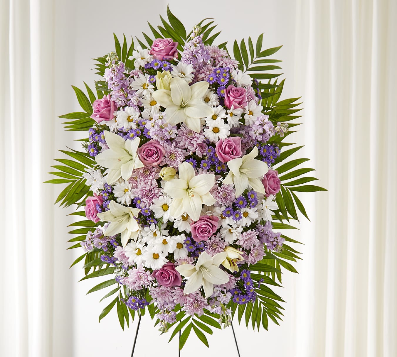 Surrounded by Love Spray - The Surrounded by Love Spray is a graceful display of remembrance, designed to honor a life with beauty and dignity. Lavender roses, stock, and cushion pompons are paired with white daisies and lilies, accented by purple monte casino and emerald palms for a lush, balanced look.  This standing spray offers a heartfelt expression of sympathy and love.  Approximately 43&quot;h x 34&quot;w
