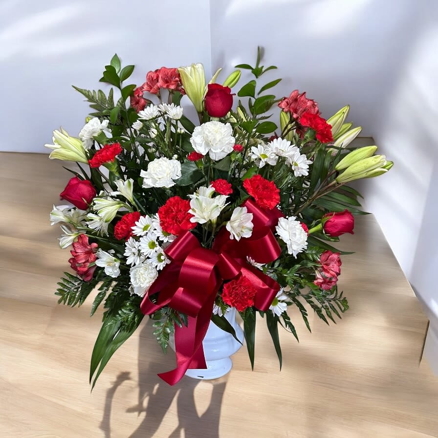 Timeless Tribute Bouquet - A heartfelt tribute arranged in a classic white urn, featuring elegant white lilies, alstroemerias, daisies, and carnations, accented with vibrant red carnations, roses, and alstroemerias. Lush palm, leather leaf, and ruscus provide a rich, textured backdrop, while a bold red bow adds a final touch of elegance and warmth