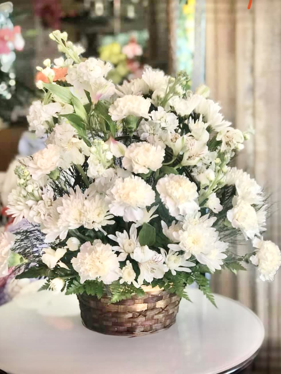 Peaceful Garden Basket - All-White Sympathy Basket Description  A serene and graceful tribute, this all-white sympathy basket exudes peace, purity, and heartfelt remembrance. Arranged in a soft woven basket—often in natural wicker or whitewashed wood—the flowers are thoughtfully layered to create a gentle, comforting presence.   Floral Composition:  White lilies take center stage, their large, trumpet-shaped blooms symbolizing the restored innocence of the soul.  White roses are nestled throughout, offering quiet elegance and representing reverence and love.  Chrysanthemums or white carnations add fullness and texture, their snowy petals evoking sincerity and devotion.  Baby’s breath or white snapdragons may be included for a delicate, airy touch, softening the arrangement with their cloud-like clusters.   Accents:  Subtle green foliage—like dusty miller or soft ferns—may peek through, adding depth without disrupting the monochromatic palette.  The basket is often lined with a satin ribbon or sheer fabric, enhancing the softness and solemnity of the display.  This arrangement is designed to convey quiet strength and compassion, offering comfort during times of loss. Add a condolence message to show your heartfelt sympathy. 