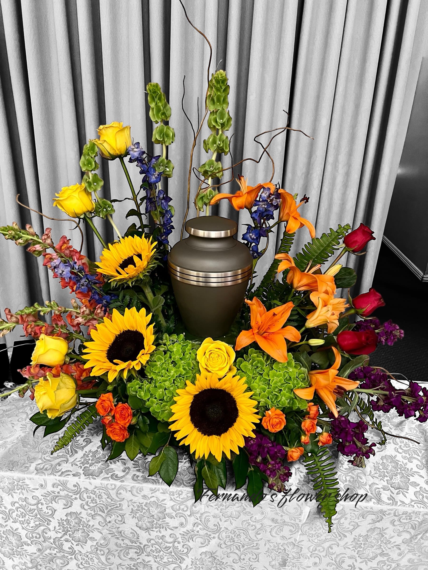 Celébrate Life - “Celebrate a life beautifully lived with this vibrant urn arrangement. ️ Featuring radiant sunflowers, bright yellow roses, striking orange lilies, and deep red blooms, this arrangement is accented with lush greenery and elegant purple and blue florals. It surrounds the urn with warmth, love, and vibrant memories, symbolizing joy and gratitude for the time shared.  Let Fernando’s Flower Shop help you create a heartfelt tribute that honors your loved one with grace and beauty. ”