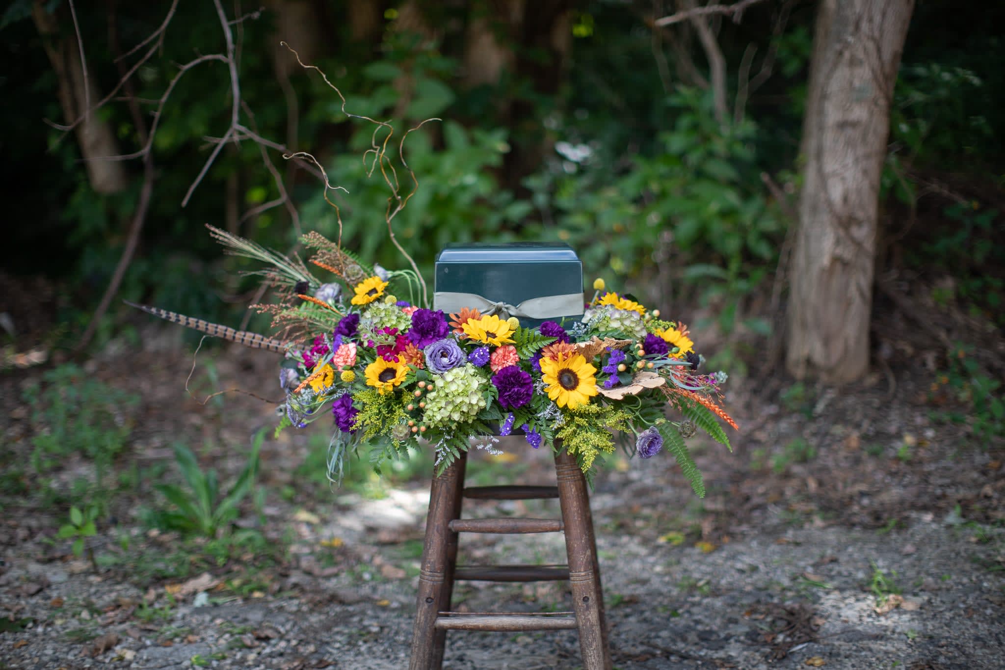Autumnal urn wreath - Seasonal funeral flowers are always a beautiful choice. Fall blooms usually include but are not limited to assorted cremons or chrysanthemums, sunflowers, pom pons, and many unusual textural elements like berries and pods of varying summer flowers. Millet, grasses and crocosmia are always a beautiful touch. Some of these blooms may be available all year long, however they are the most striking late August through November. Please call if ordering off season. Customized script is available for all floral tributes. Please include urn dimensions in special instructions. 