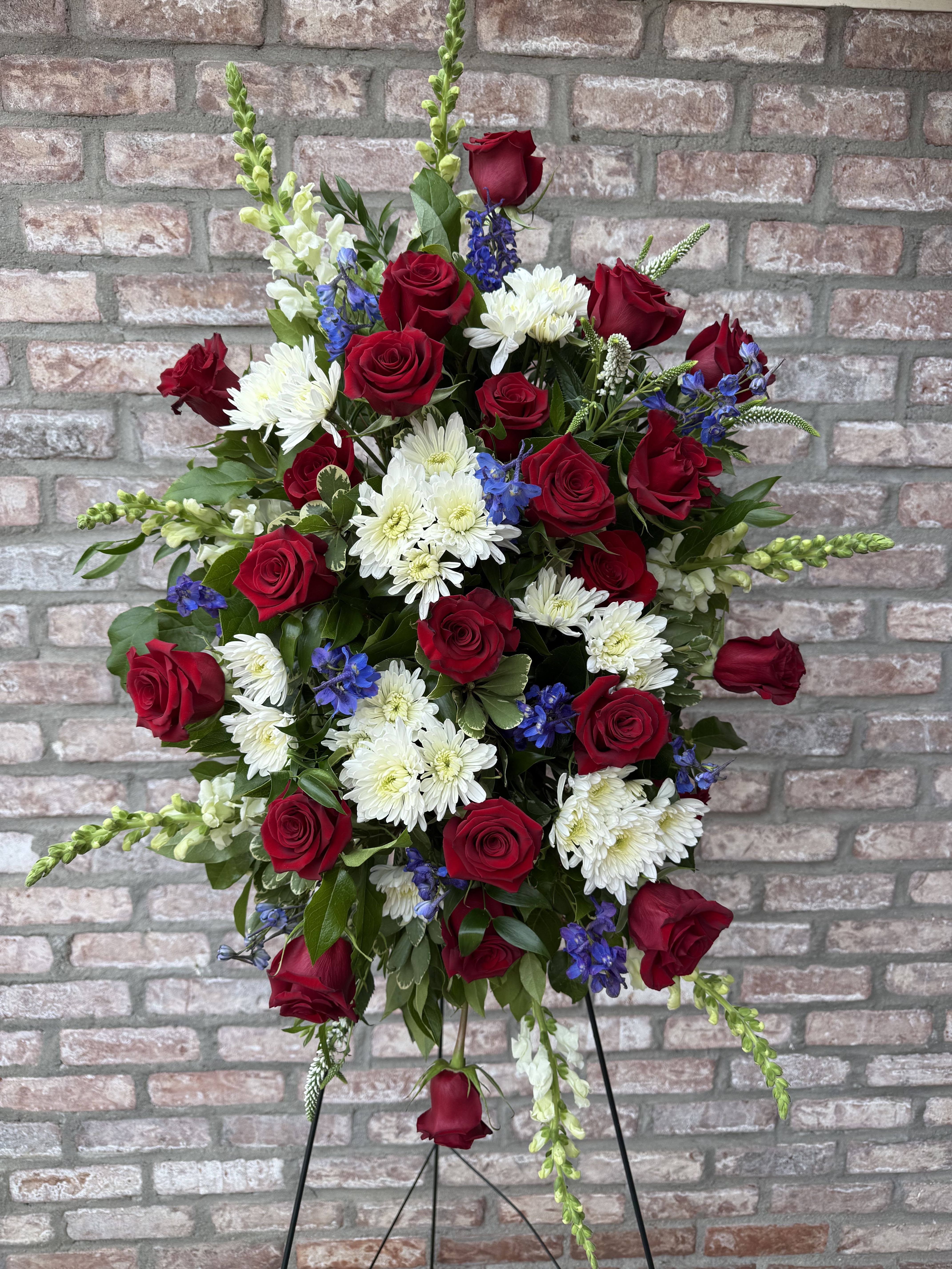 Peaceful Warrior Easel Spray by Simply Flowers - A Red, White and Blue easel tribute filled with red roses, white chrysanthemums, white snapdragon, and blue delphinium. Measures approximately 30” tall and 24” wide. Comes with a 60” easel. 