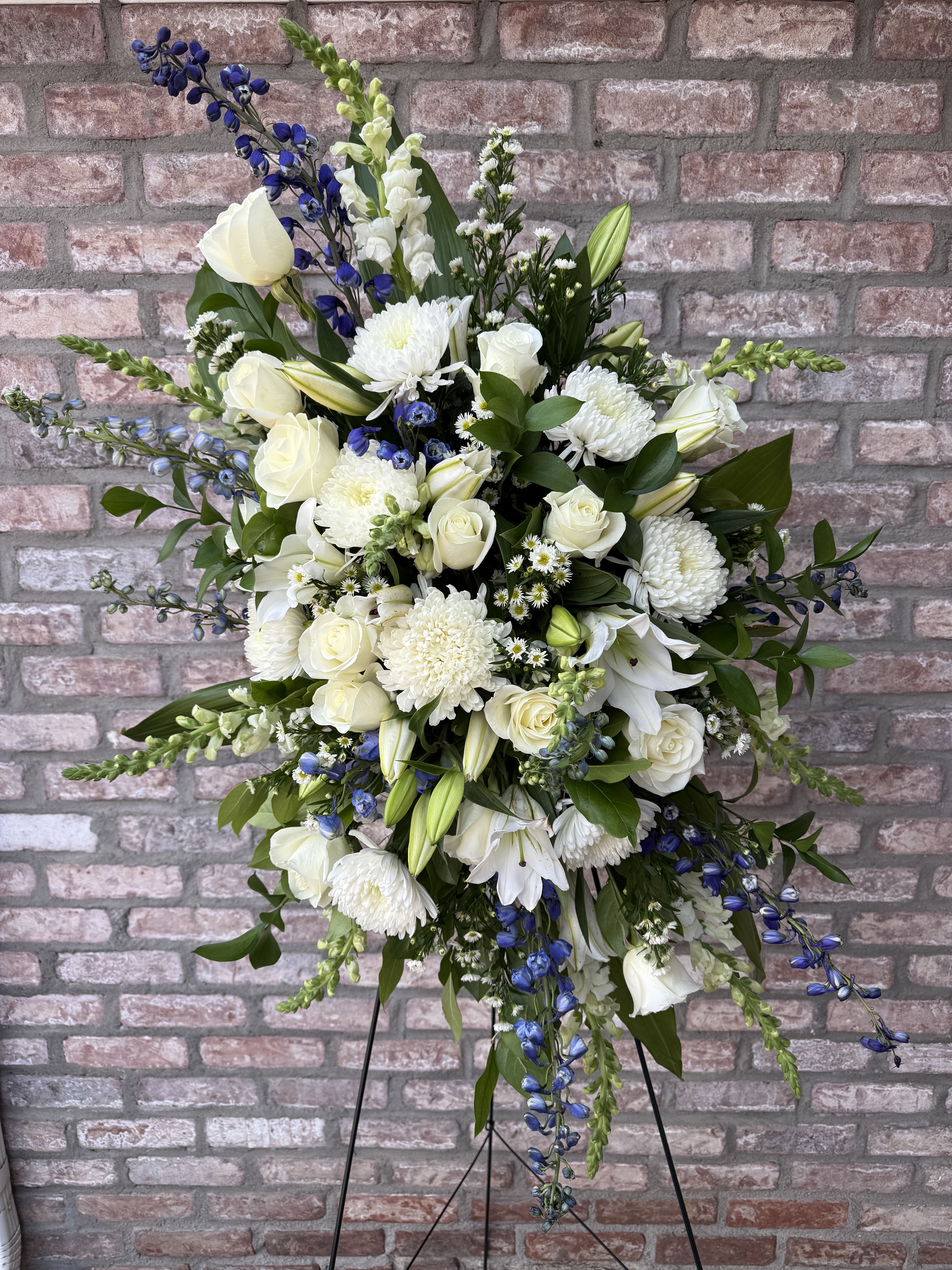 Peaceful Remembrance Easel Spray by Simply Flowers - A fragrant, lush design of white Oriental lilies, white roses, snapdragon, delphinium and more creates this beautiful easel tribute exuding peace and comfort. Measures approximately 36” tall x 28” wide. Comes with a 60” easel. 