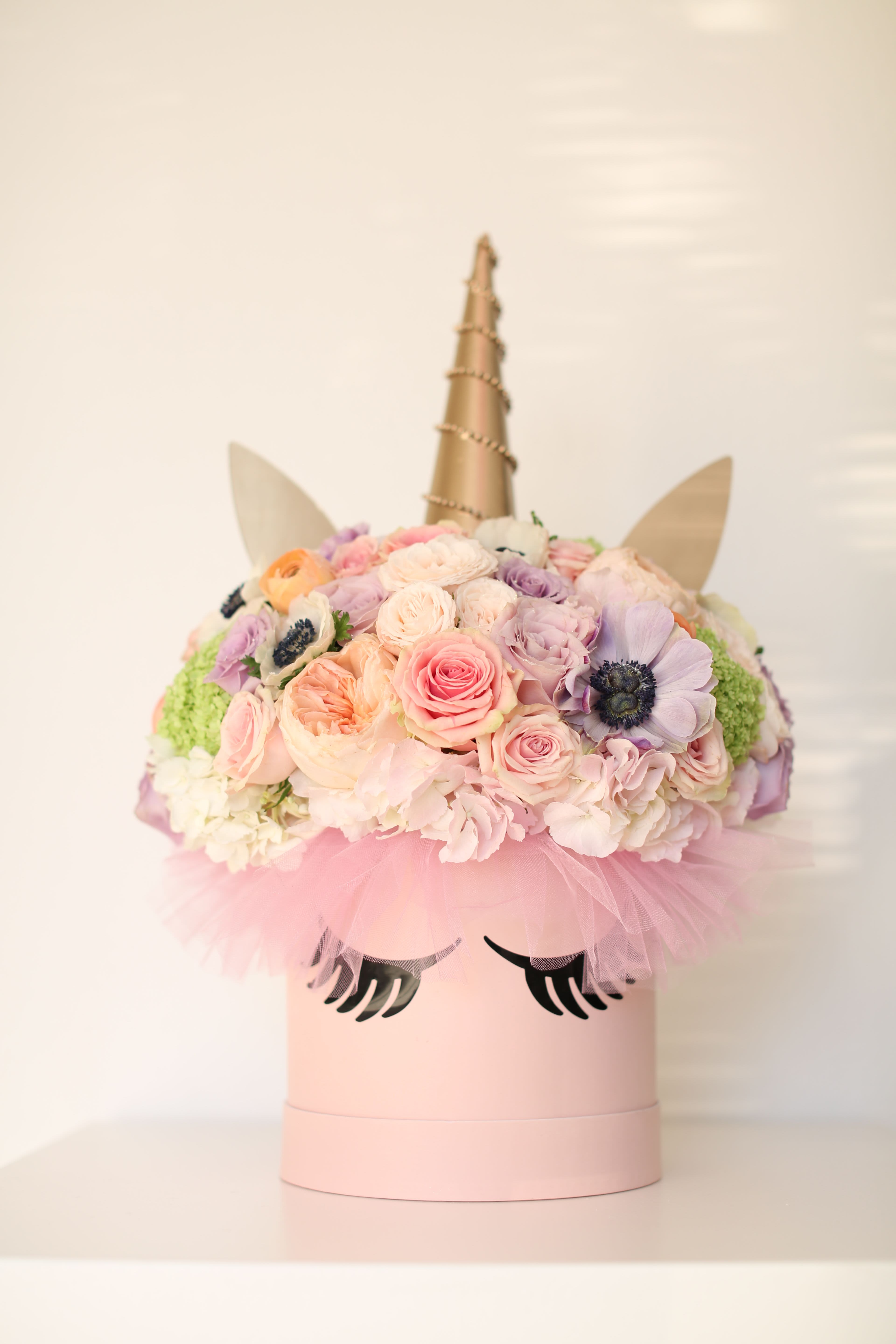 Enchanted - Make every day magical with this whimsical unicorn arrangement that brings fairy tale dreams to life! This adorable pink hatbox features sweet eyelashes and a shimmery golden horn, transformed into the most enchanting floral unicorn you've ever seen.  A cloud of pastel blooms creates the unicorn's mystical mane, featuring soft pink roses, lavender roses, and creamy ranunculus that seem to shimmer like unicorn magic. Delicate anemones with dark centers add contrast and charm, while bright green button mums provide pops of fresh color. Peachy garden roses and blush spray roses complete this dreamy palette that's as soft and sweet as cotton candy.  The arrangement is finished with a fluffy pink tulle collar that adds the perfect touch of whimsy, making this unicorn ready to prance into any celebration. The golden horn is wrapped with delicate pearl garland, adding an extra touch of sparkle and elegance.  Perfect for: - Little girls' birthdays and parties - Baby showers and nursery décor - First communions and special milestones - Anyone who believes in magic - Unicorn-themed celebrations - &quot;Just because&quot; smiles   - Approximately 12-14 inches tall 11 inches wide  Where flowers meet fairy tales – this unicorn bouquet proves that magic is real and it smells absolutely beautiful!