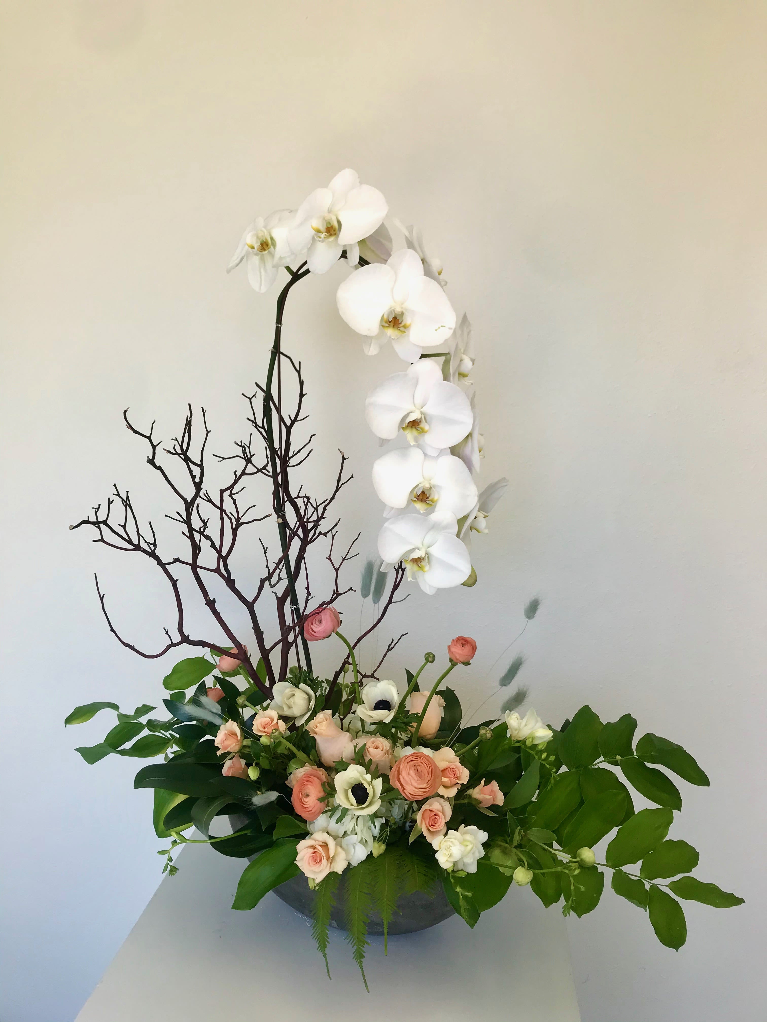 Enchanted Orchid Garden - &quot;Some flowers whisper. This arrangement speaks poetry.&quot;  This breathtaking arrangement captures the essence of refined beauty with its graceful cascade of a pristine white phalaenopsis orchid dancing above a garden of delicate roses,  ranunculus, and a bed of lush emerald foliage  Thoughtfully designed with an artistic manzanita branch that add sculptural drama, this arrangement tells a story of seasons - the promise of spring's renewal intertwined with nature's quiet strength. The contrast between the smooth orchid petals and the textured branches creates visual poetry that transforms any space into a sanctuary of calm sophistication.  Perfect For: - Luxury home decor that speaks to discerning taste - Corporate reception areas seeking understated elegance   - Special celebrations where beauty matters most - Sympathy expressions offering comfort through natural grace - Anniversary gifts symbolizing enduring love and growth  - Height: Approximately 24-26 inches   