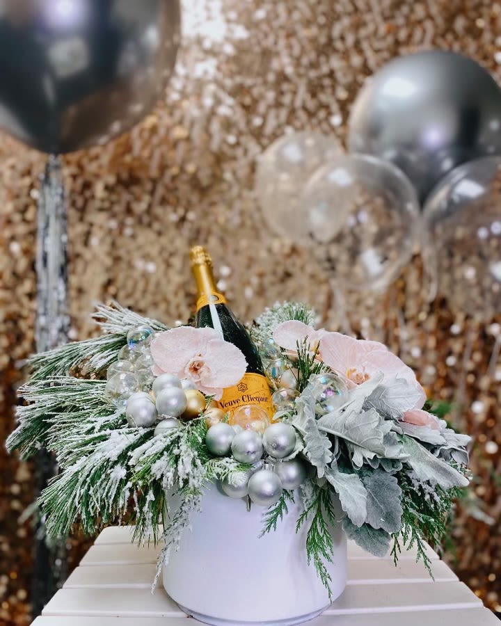 Jubilee - Celebrate in style with this show-stopping arrangement that combines the sophistication of premium Veuve Clicquot champagne with the natural beauty of winter florals. Presented in a sleek white ceramic container, this arrangement strikes the perfect balance between festive celebration and elegant sophistication. The champagne bottle nestled among winter florals creates an unexpected and delightful surprise that guests will absolutely adore. It's luxury you can see, smell, and eventually taste!  Perfect For Life's Celebrations - New Year's Eve parties that demand glamour and style - Holiday entertaining when you want to impress - Anniversary celebrations marking life's golden moments - Corporate holiday gifts that make a lasting impression - Housewarming present for the host who has everything - Engagement parties celebrating love and new beginnings - Milestone birthdays deserving of champagne toasts - Holiday hostess gifts that show true appreciation  Dimensions: Approximately 14&quot; wide x 10&quot; tall   Includes: 750ml Veuve Clicquot bottle   Perfect for: Ages 21+ celebrations  