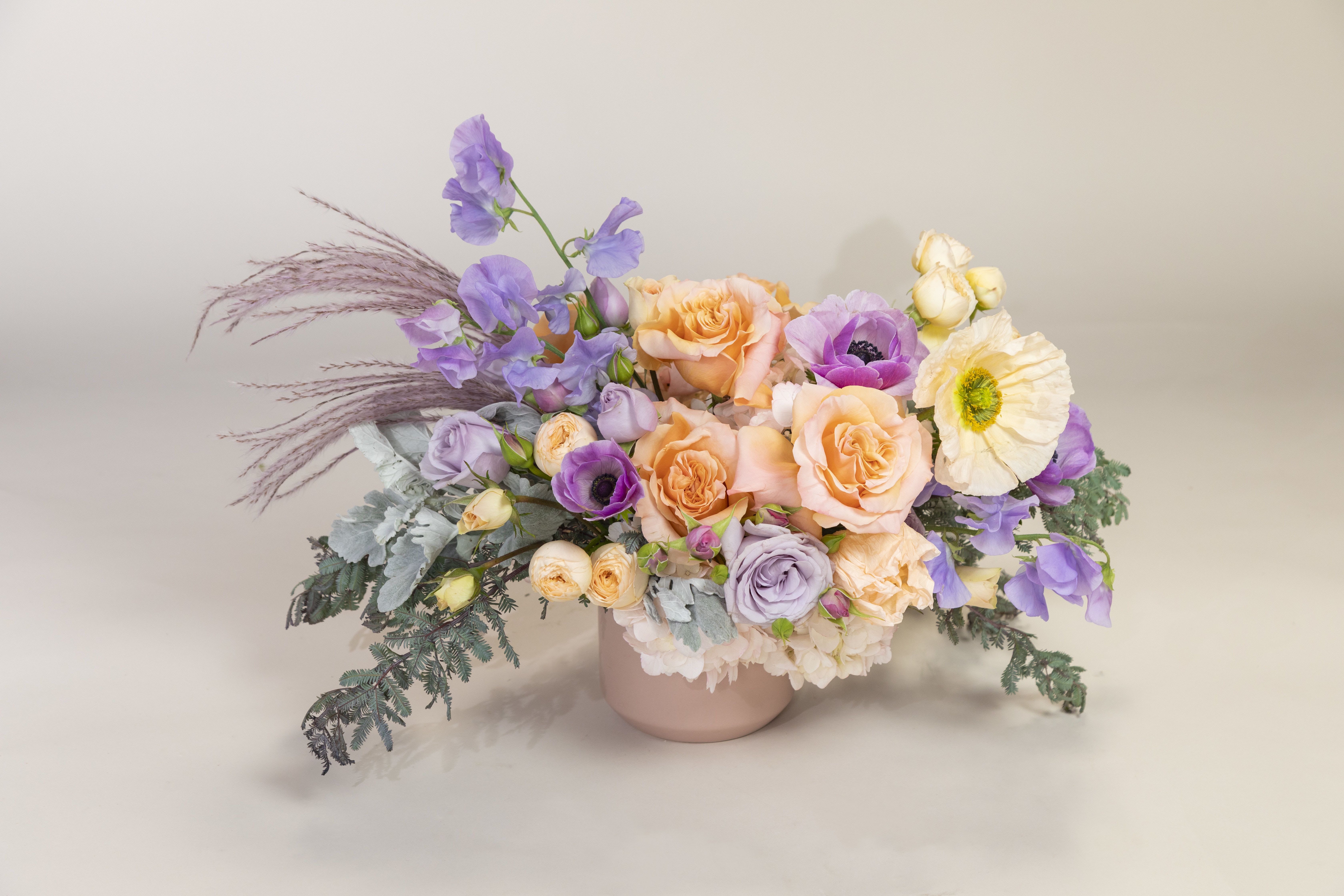 Juliet's Charm - This bestseller is made up of blush hydrangea, lavender anemones, peach roses, lavender roses, lavender sweet peas, dramatic Icelandic poppies, accented with lavender stypha set inside a pink 5&quot;x5&quot; ceramic vase.