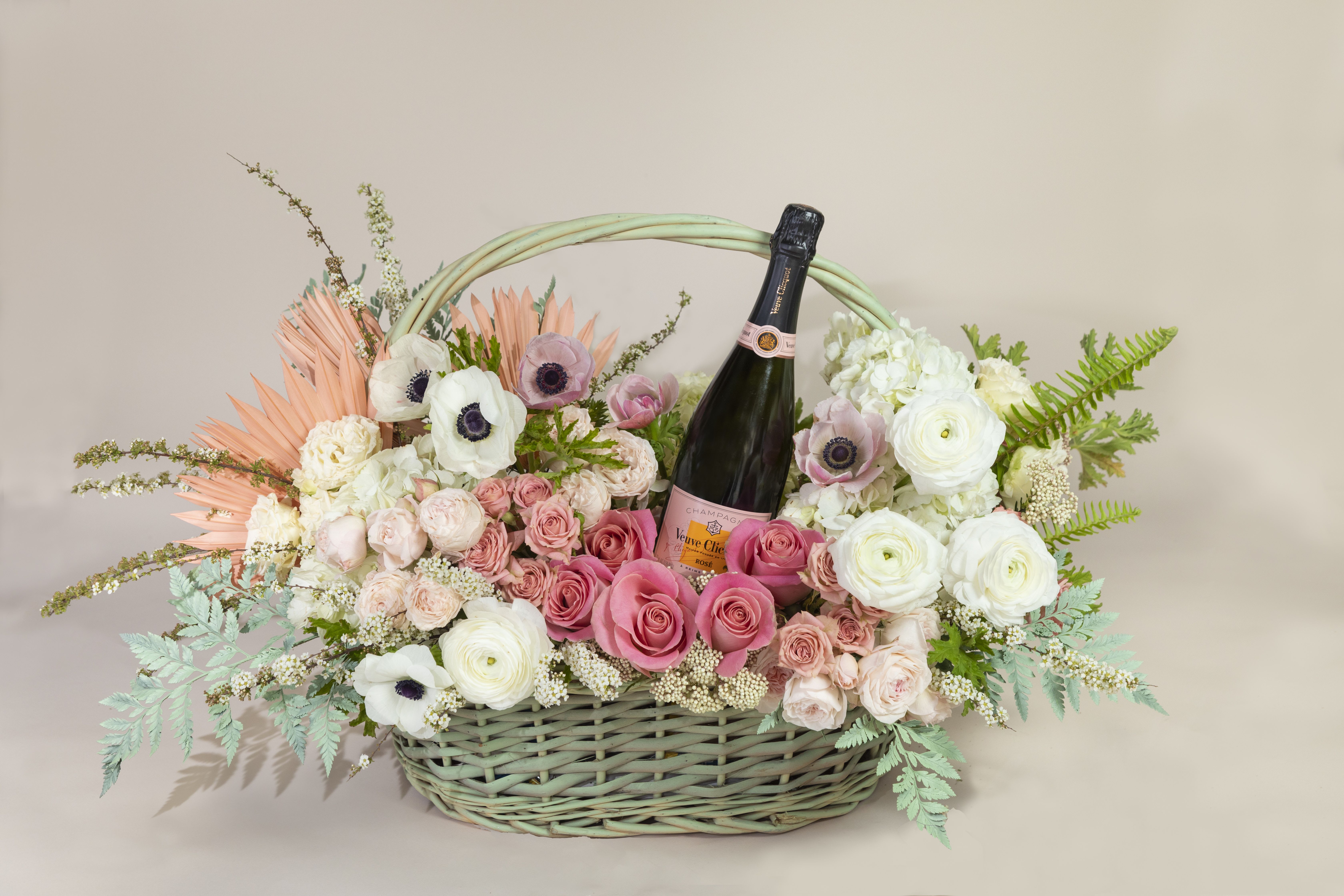 Sunrise with Mimosas - This extra large floral arrangement is made inside an 18&quot; Basket with a premium bottle of Veuve Clicquot nestled in upscale blooms that include roses, spray garden roses, classic spray roses, anemones, ranunculus, hydrangeas. Accented with dried palm fans