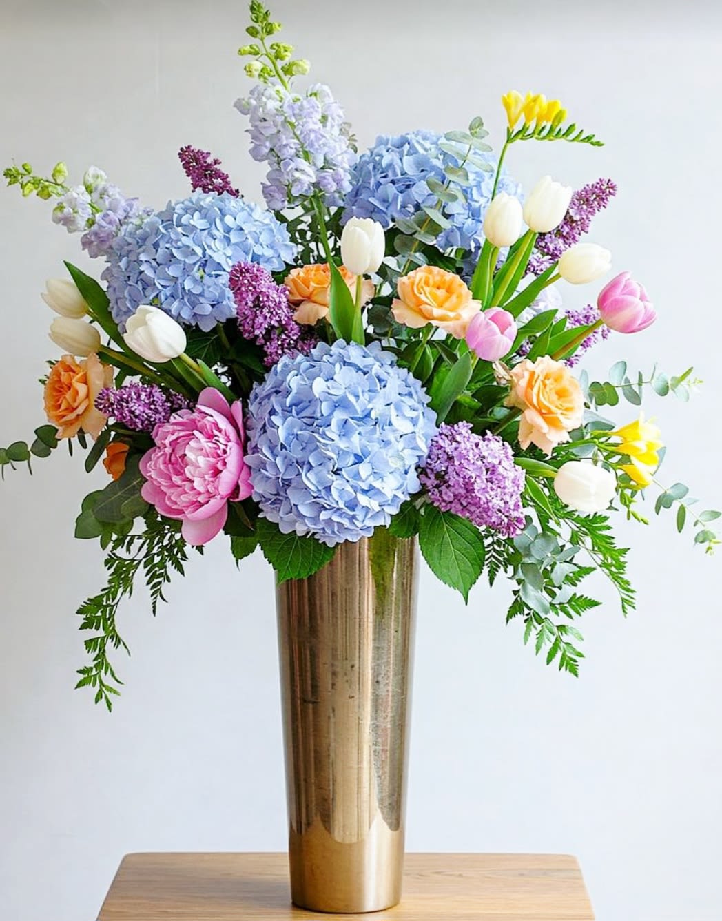 Goldie by Pretty in Petals  - This arrangement is designed to bring a touch of sunshine and elegance to any space, perfect for celebrating special occasions or adding a bright, cheerful accent to your home. The combination of rich yellows, purple, and complementary blue, paired with a gold vase, creates a warm and inviting display.