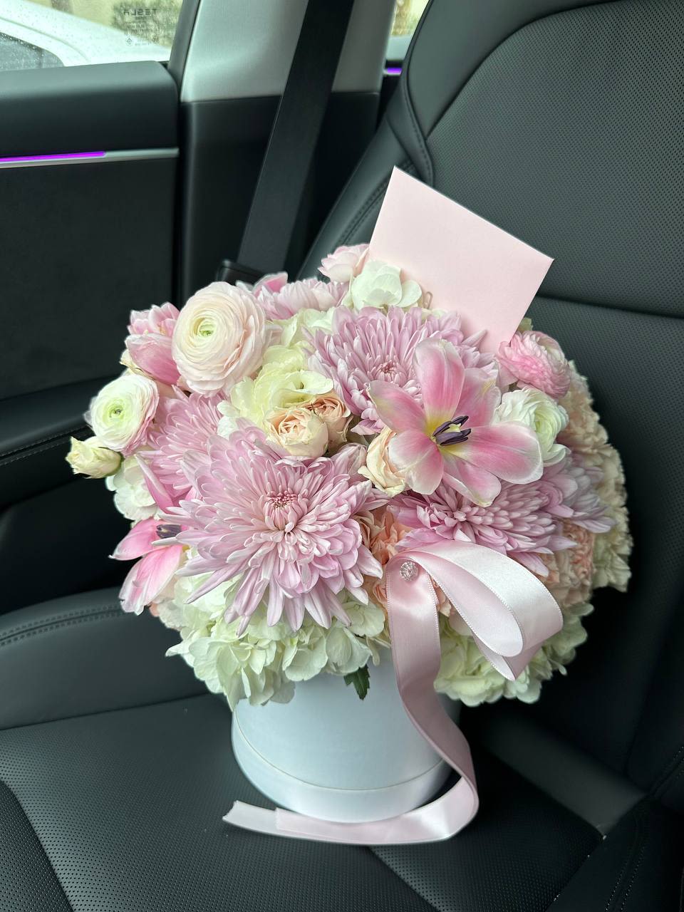 Tender Bloom - Tender Bloom is an exquisite floral arrangement that combines the lushness of hydrangeas, the delicate charm of ranunculuses, the classic beauty of roses, the subtle elegance of carnations, and the fresh appeal of tulips. These flowers are thoughtfully arranged to create a bouquet that embodies elegance and romance.  The hydrangeas add volume and a sense of luxury with their large, cloud-like blossoms. Ranunculuses, known for their layered petals, contribute texture and grace. Roses, timeless symbols of love and devotion, infuse the arrangement with classic romanticism. Carnations, with their ruffled petals, bring softness and sophistication. Tulips, representing spring renewal, introduce a touch of freshness and lightness.  Presented in a stylish hatbox and adorned with a satin ribbon, Tender Bloom offers a modern and elegant aesthetic. This arrangement is perfect for special occasions, whether it’s a declaration of love, an expression of gratitude, or simply a desire to bring joy to someone dear.