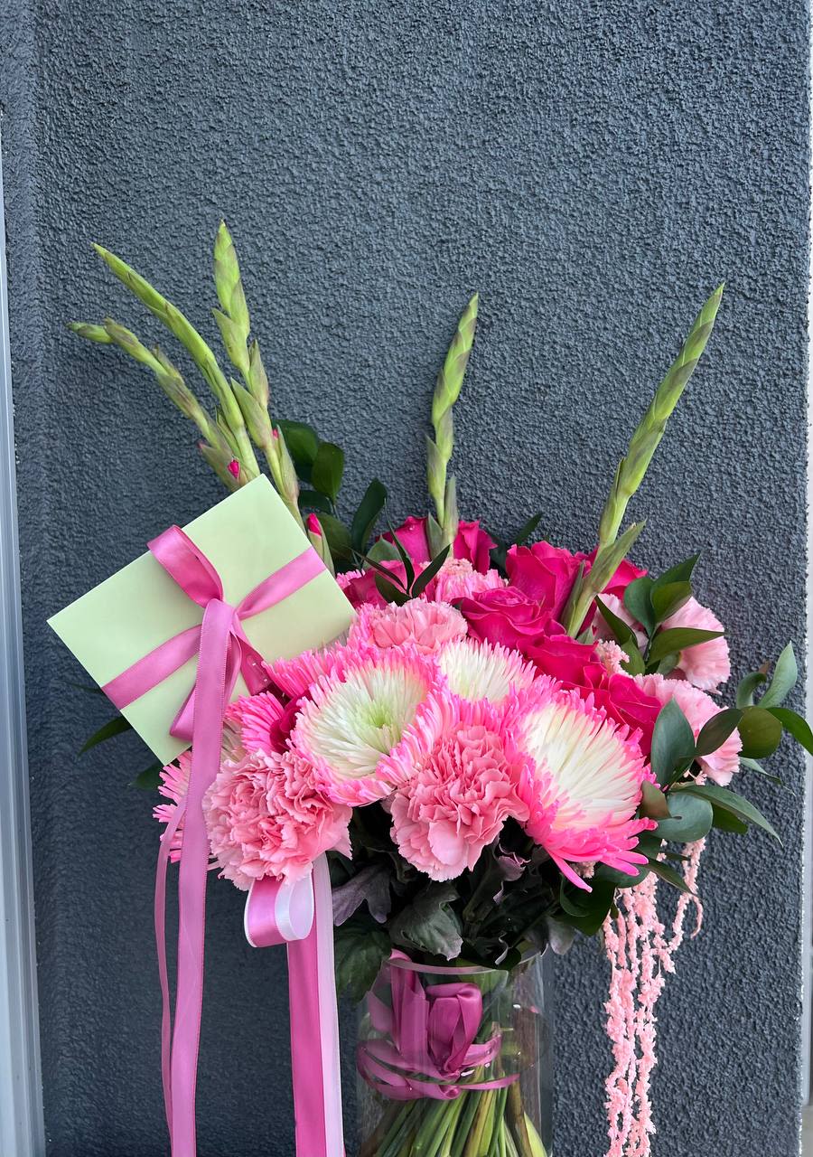 Flamingo Bliss Flamingo Bliss - Flamingo Bliss is a vibrant and stylish bouquet that radiates energy, passion, and elegance. Its rich pink hues, complemented by soft green accents, evoke the grace of flamingos basking in the warmth of the tropical sun. This arrangement features exquisite roses, airy carnations, striking chrysanthemums, and elegant gladioli, creating a sense of lightness, playfulness, and sophistication. The accent ribbons and chic presentation make this bouquet truly special. THE BOUQUET COMES WITH A VASE