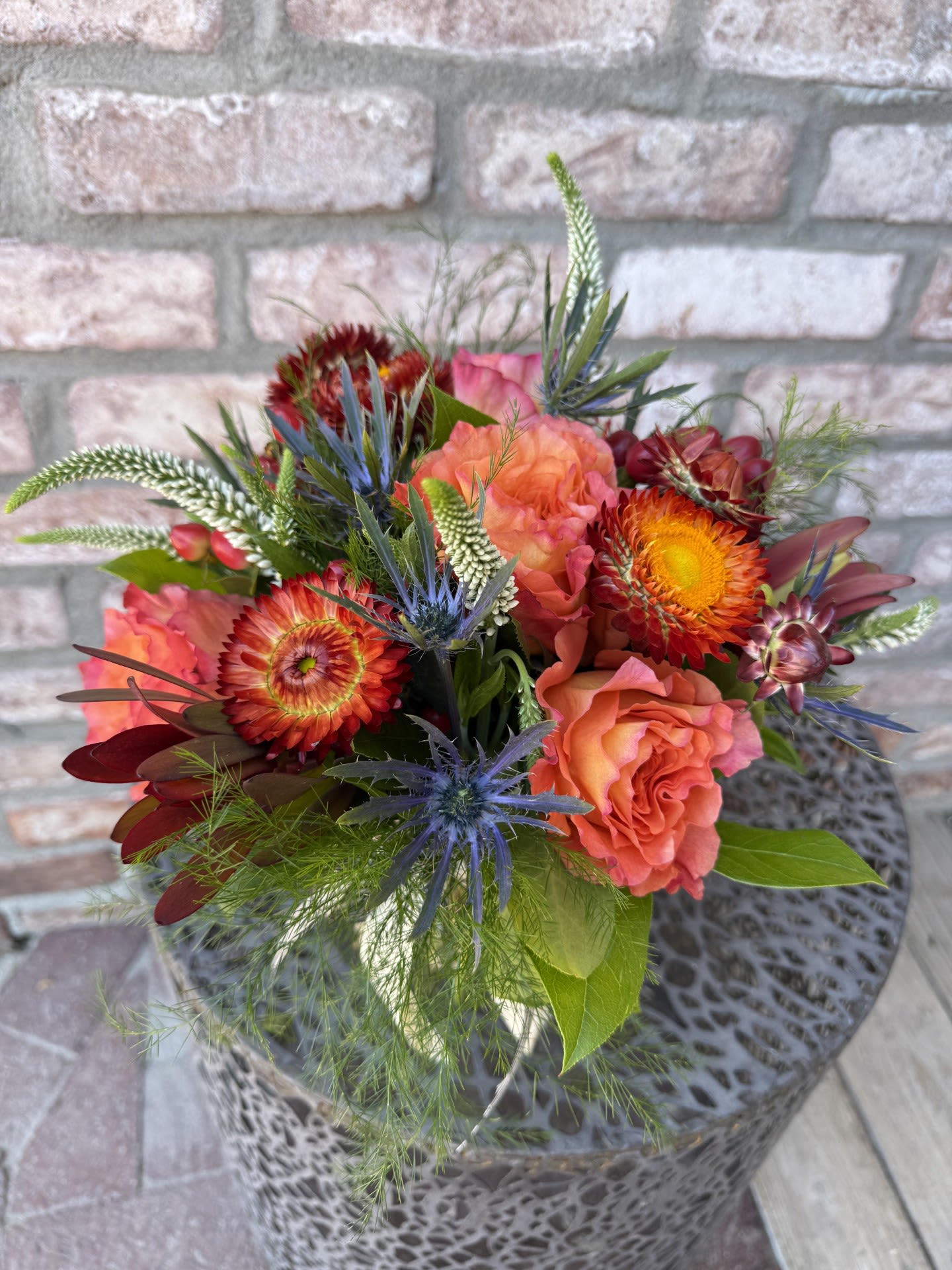 New! Harvest Glow - Designed in a glass vase, filled with lush greenery and beautiful pops of color! Roses, strawflower, eryngium, veronica and more. Perfect for a side table or desk. Measures approximately 10&quot; tall.